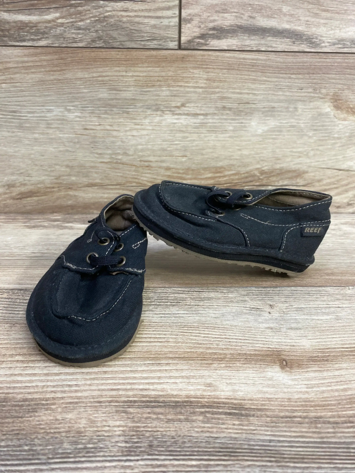 Three-quarter view of Reef Grom Deckhand 3 toddler sneakers in black canvas showcasing moc-toe stitching and elastic laces on a light wood floor