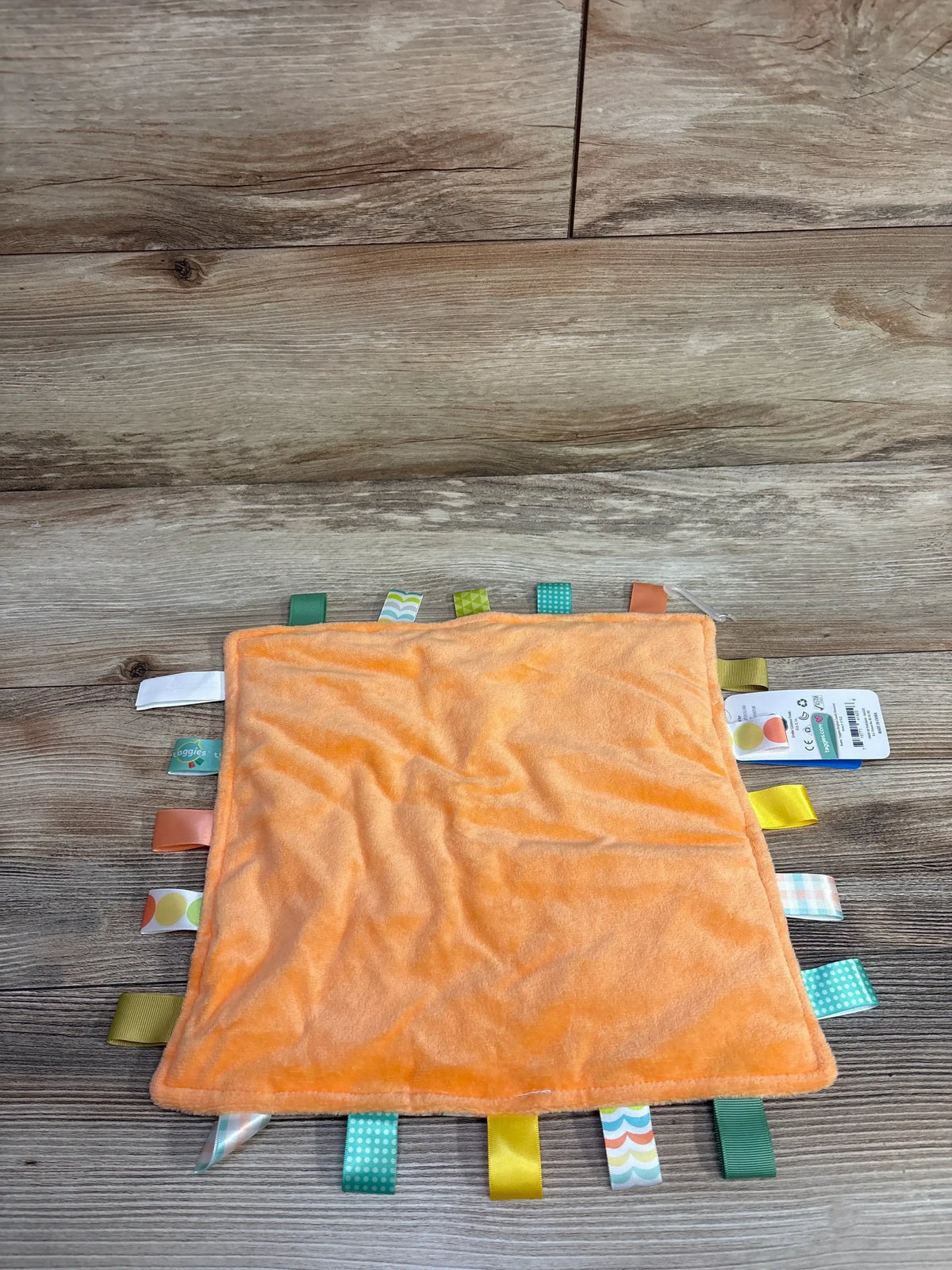 Top-down view of Taggies Original Comfy Dinosaur Blanket in soft orange showcasing colorful ribbon sensory tags on wood background