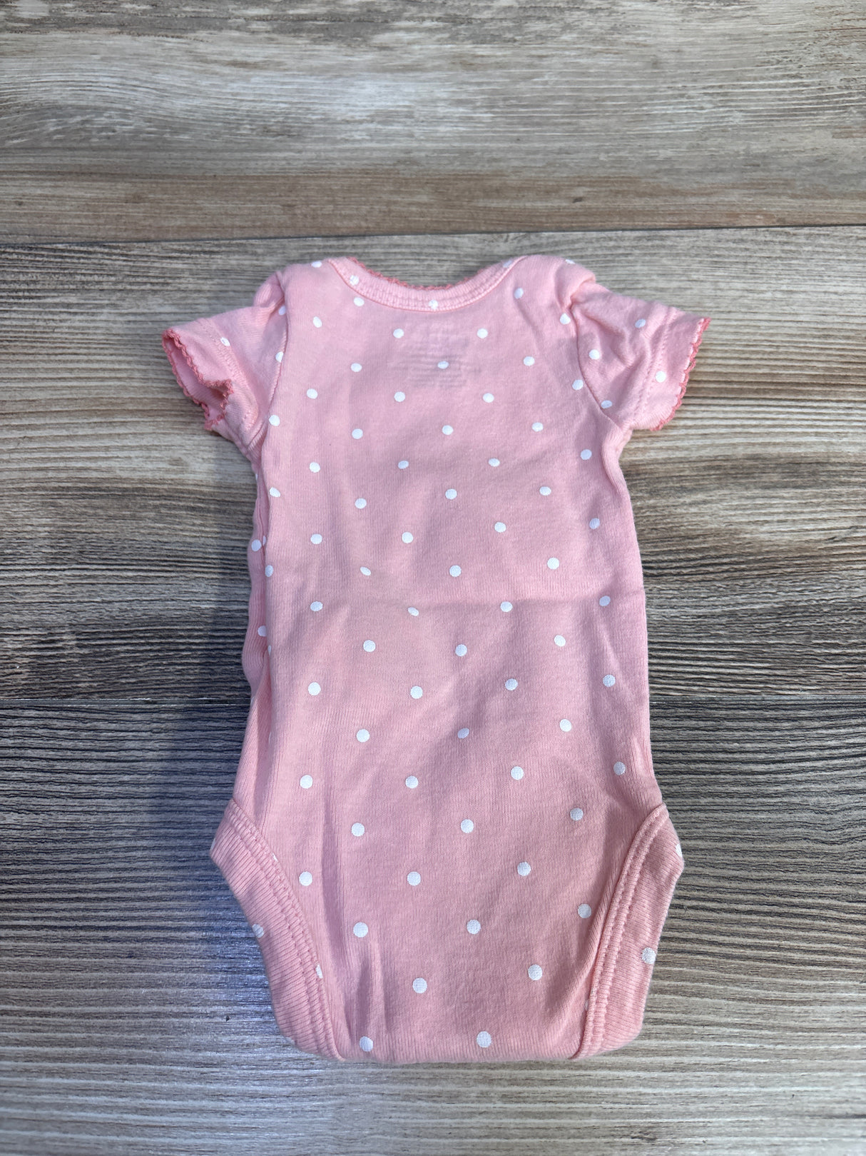Flat lay of Simple Joys pink preemie bodysuit showing white polka dot pattern and short ruffled sleeves on wood background