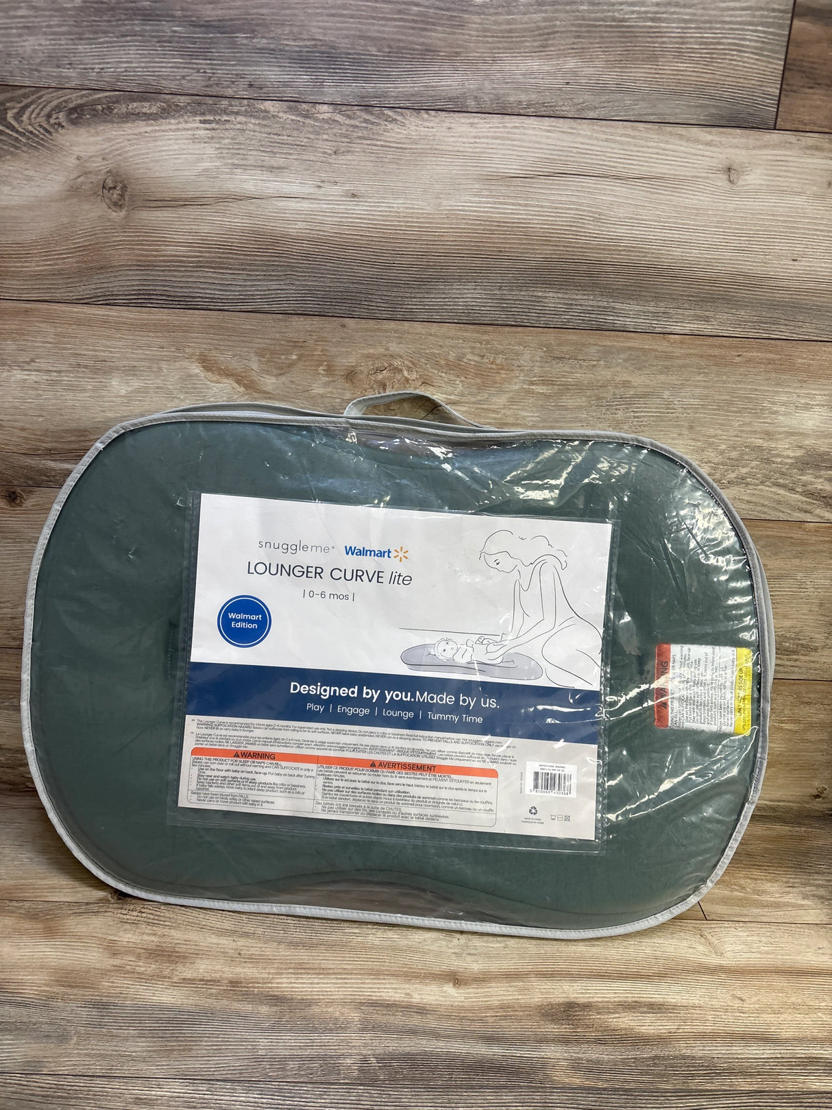 Top-down view of Snuggle Me Curve Lite baby lounger with moss green cover in clear plastic packaging on wood floor