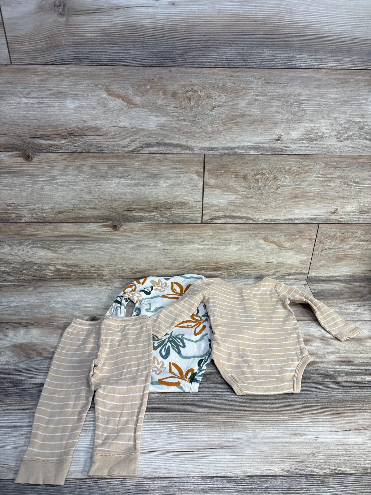 Overhead view of Carter’s beige three-piece bodysuit set showcasing two bodysuits and pants with a wood floor background