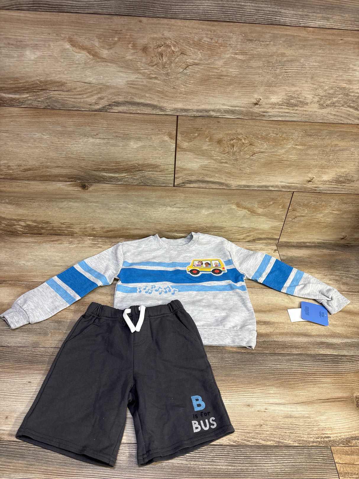 Flat lay of toddler grey sweatshirt with blue stripes and school bus patch paired with black drawstring shorts reading B is for BUS on wood floor, size 4T
