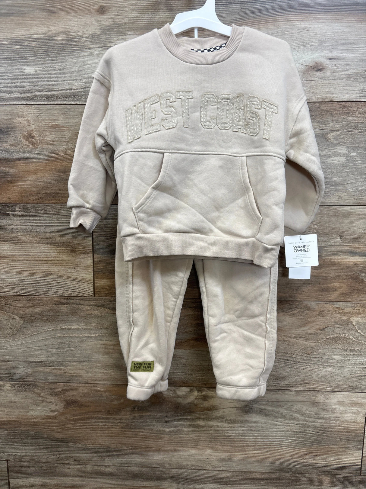 Front view of beige fleece crewneck and jogger set with WEST COAST applique, on white hanger against wood background