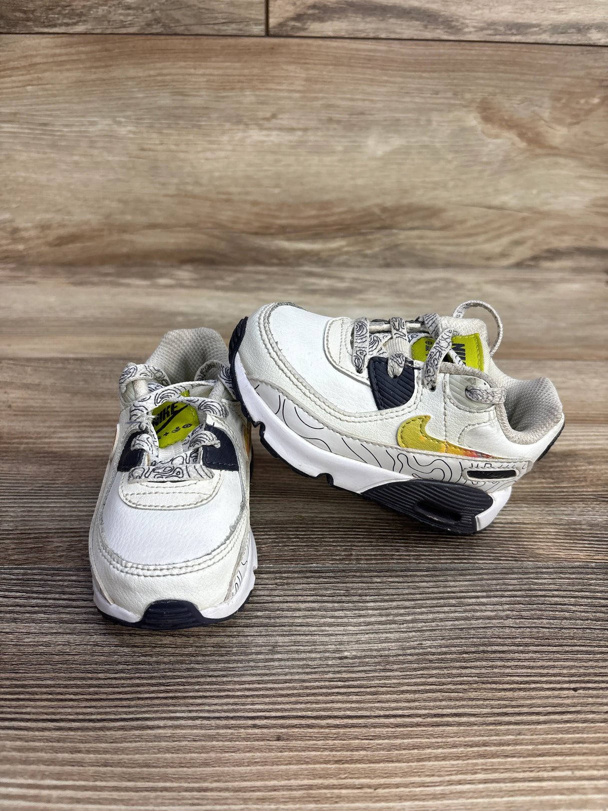 Pair of white Nike toddler sneakers with yellow swoosh, black sole, and patterned laces on wooden floor