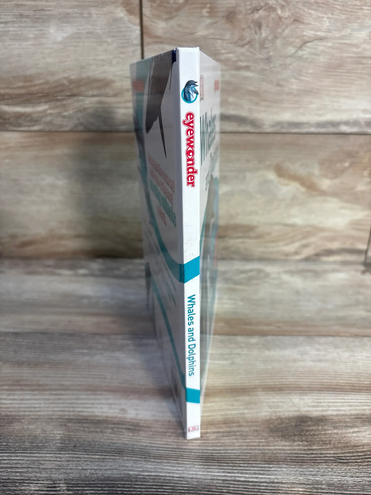 Side view of Eye Wonder Whales and Dolphins Hardcover Book showcasing its spine on a wood grain background