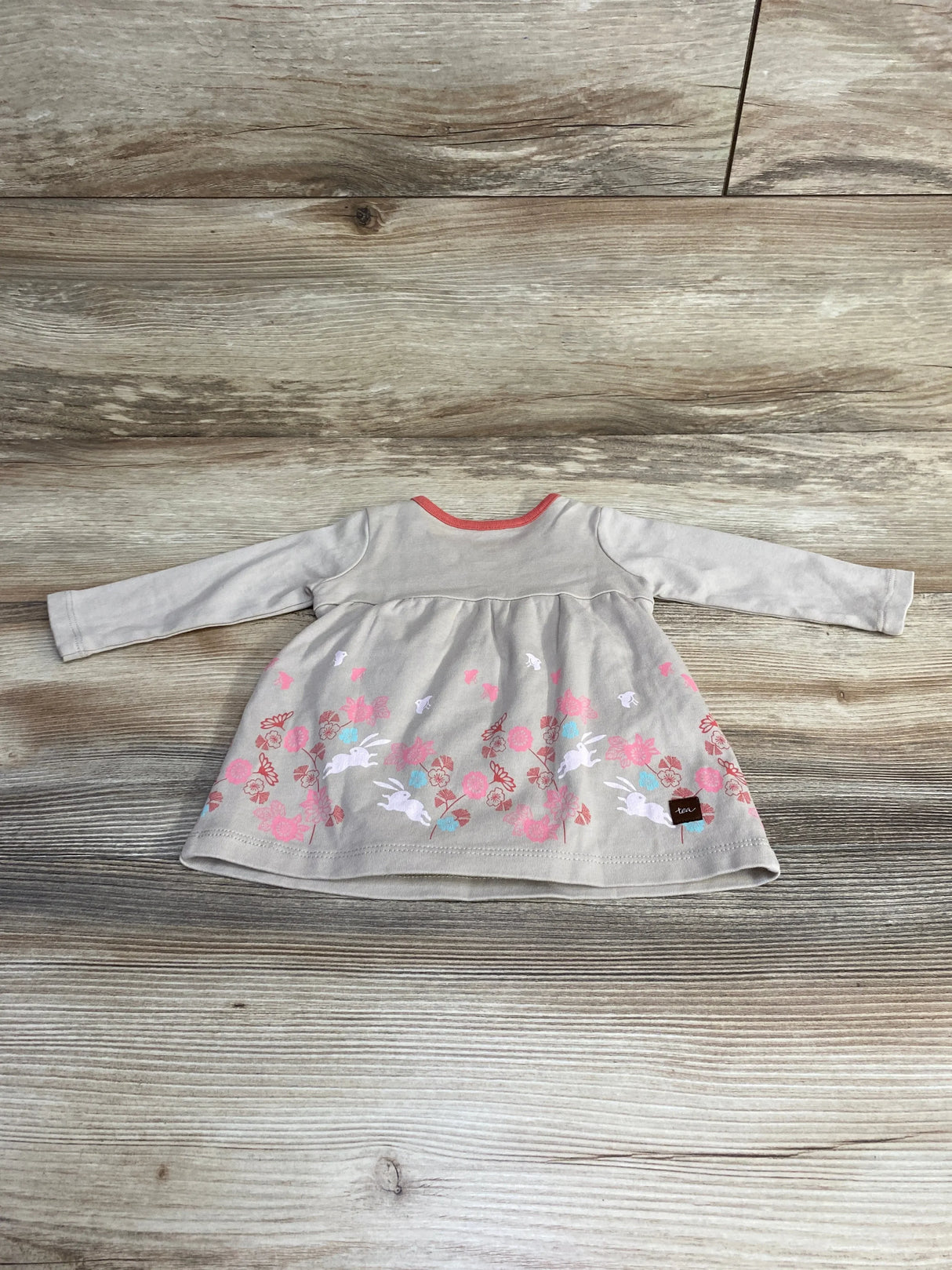 Front view of newborn beige long-sleeve dress with coral neckline and pink floral-and-bunny border print, laid flat on wood background
