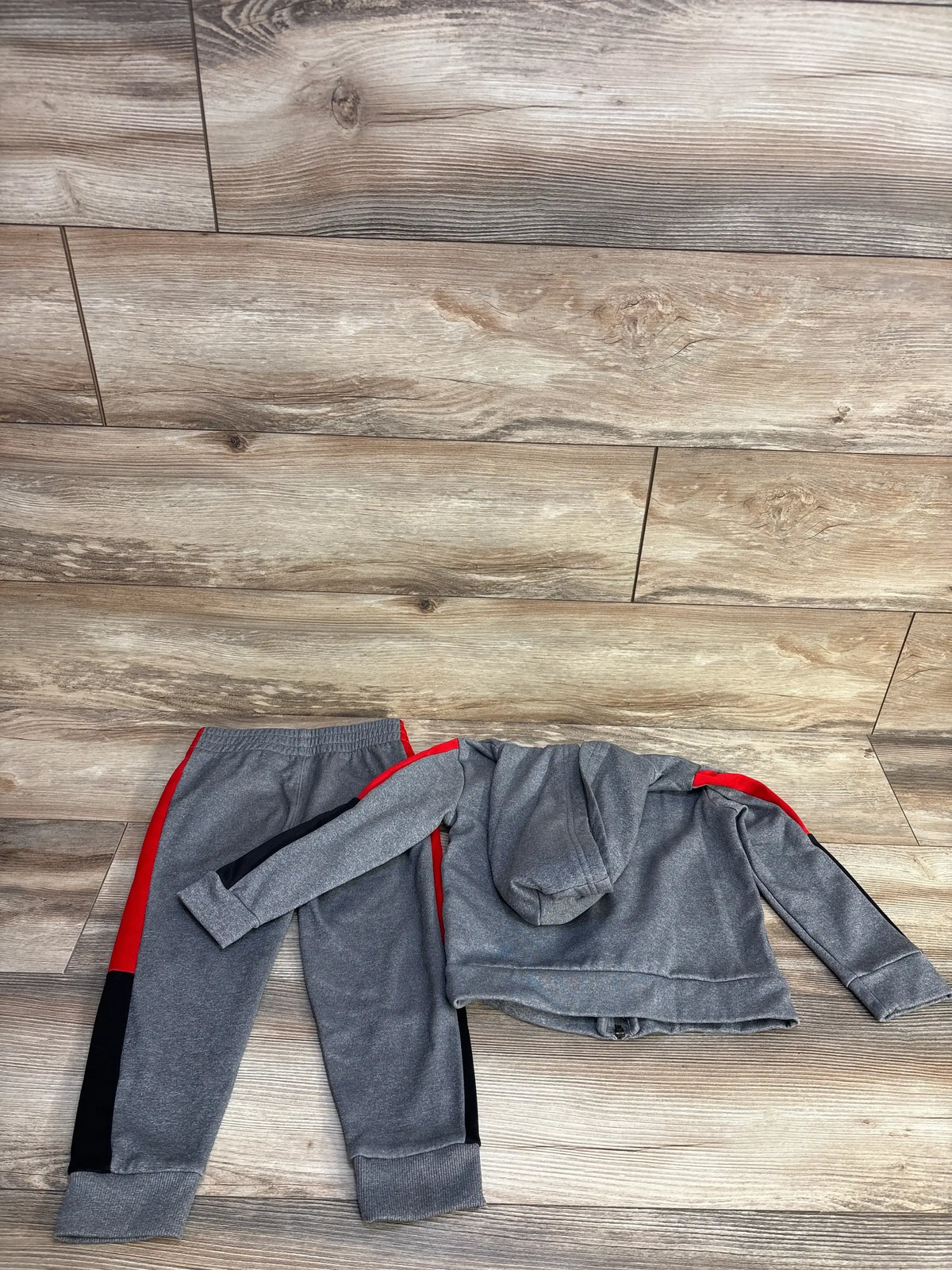 Flat lay of grey Puma 2pc tracksuit size 4T showing hooded full zip jacket and pants with red and black side stripes on wood floor