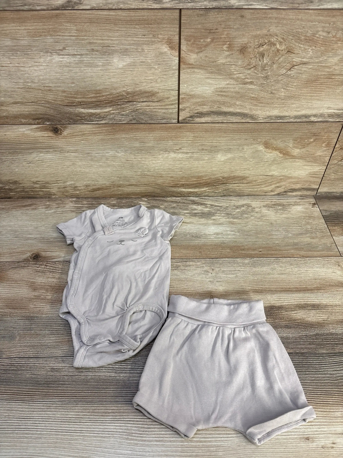 Top-down flat lay of beige kimono-style bear bodysuit with side snaps and matching foldover shorts on wood background