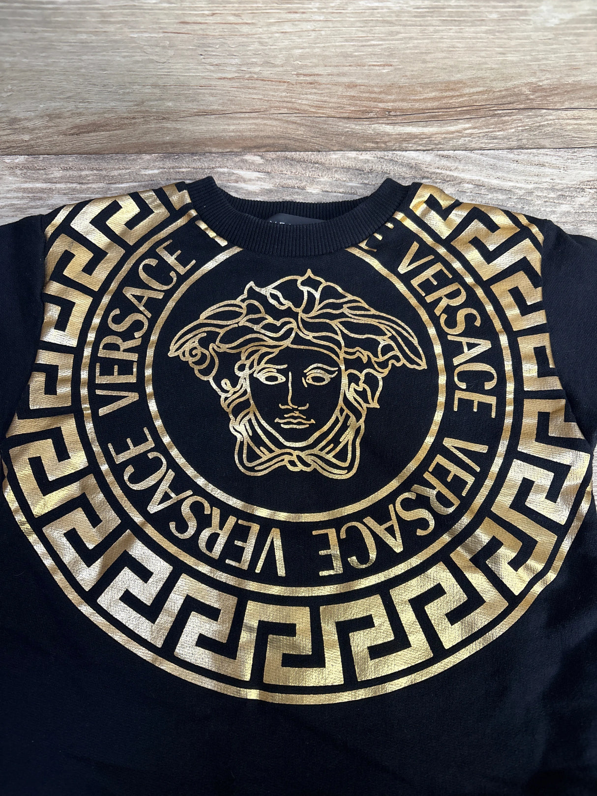 Black Versace shirt with gold Medusa logo and Greek key pattern on wood background