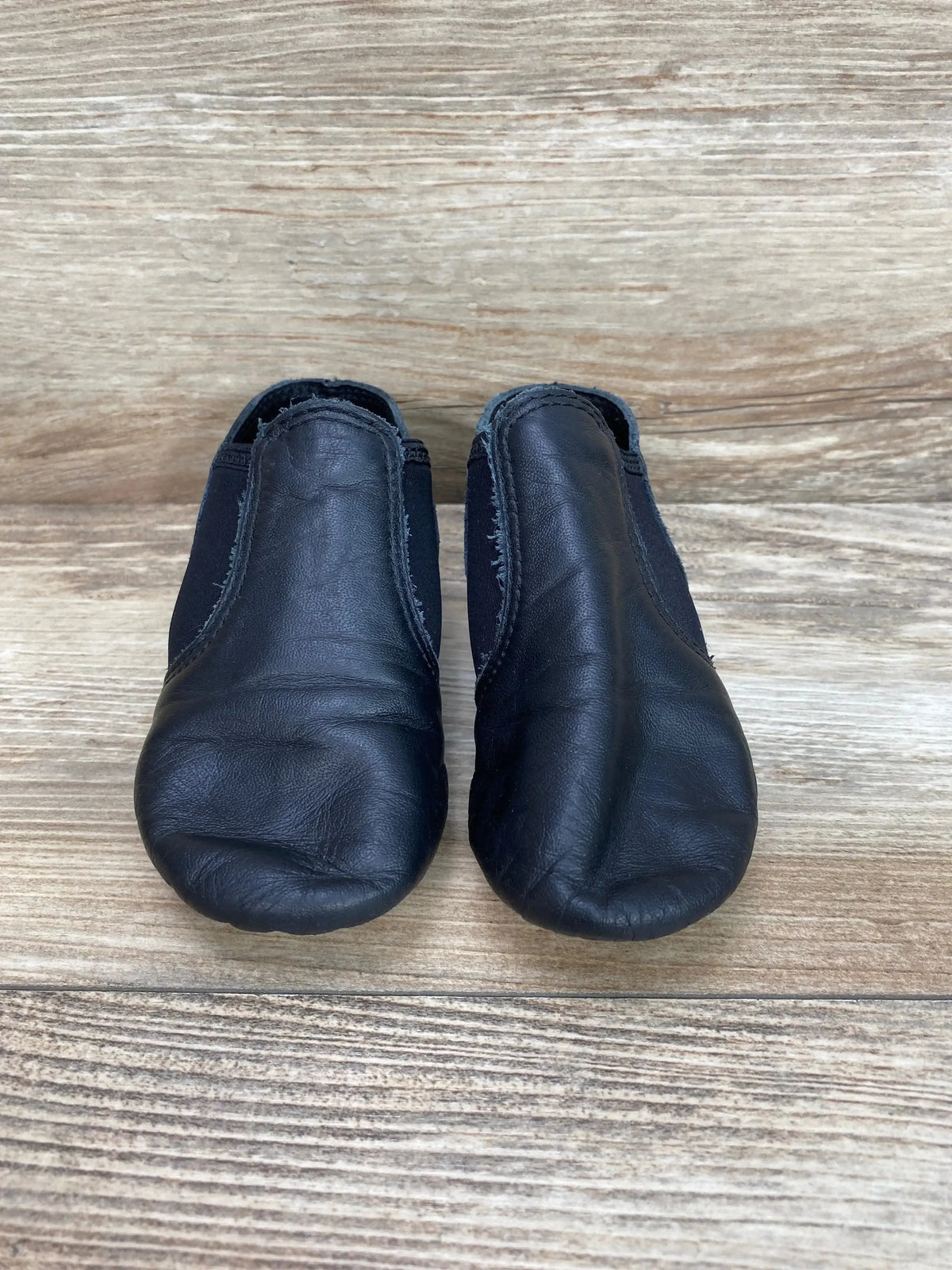 Front view of Capezio black jazz slip-on shoes, child size 10C, pair showing elastic sides and worn toes on wood background