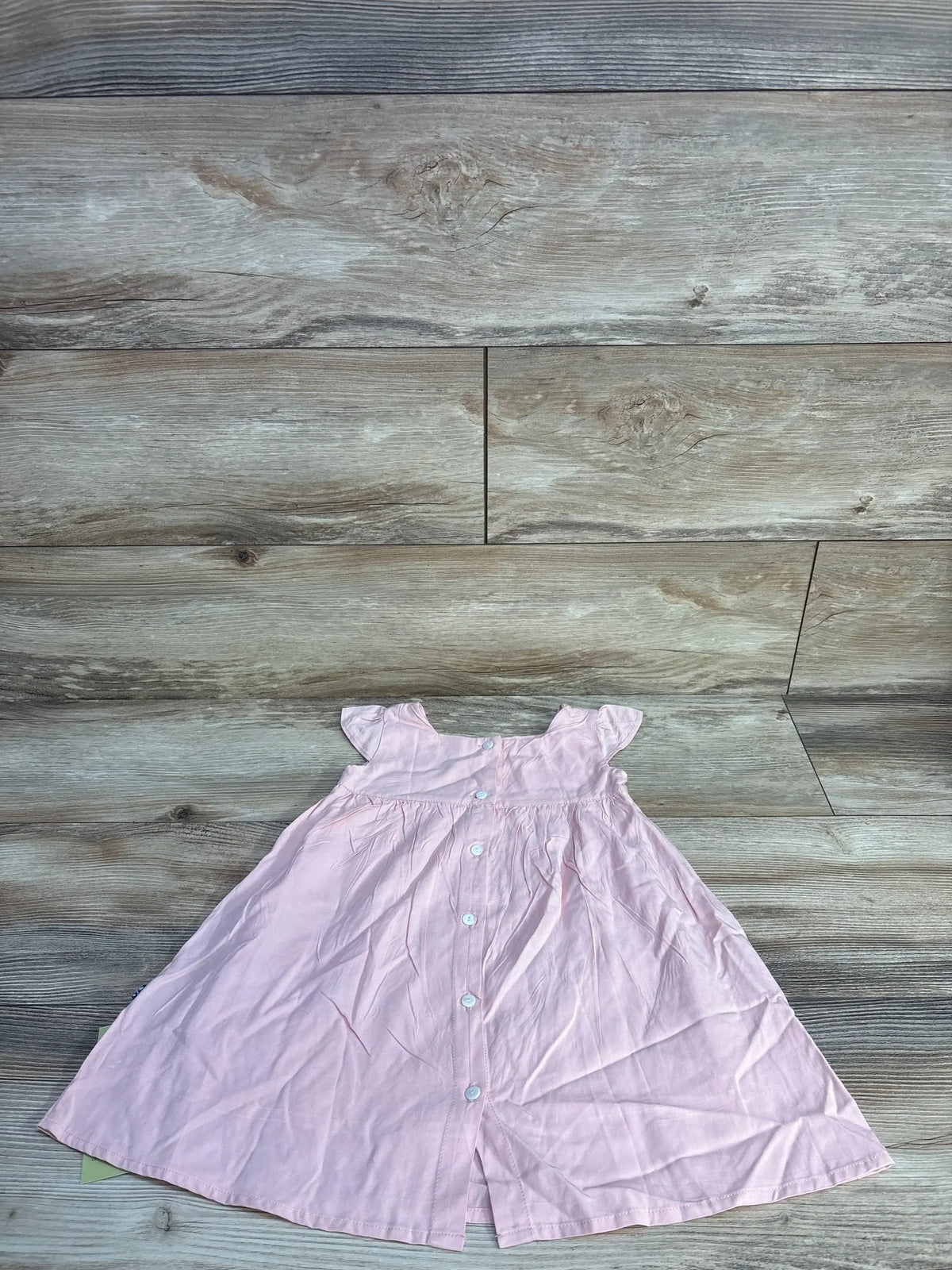 Flat lay of pink woven toddler dress size 12-18 months showcasing apron-style bodice and front buttons on wood background