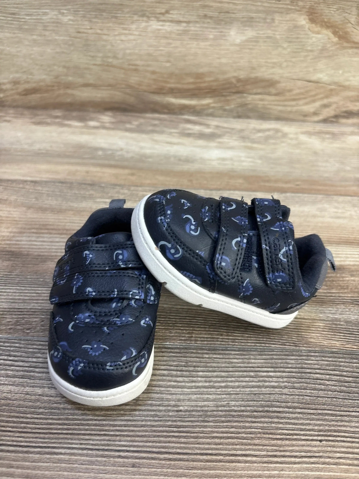 Top view of Carter’s Neo Dino navy sneakers showcasing two hook-and-loop straps and white soles on a wood surface