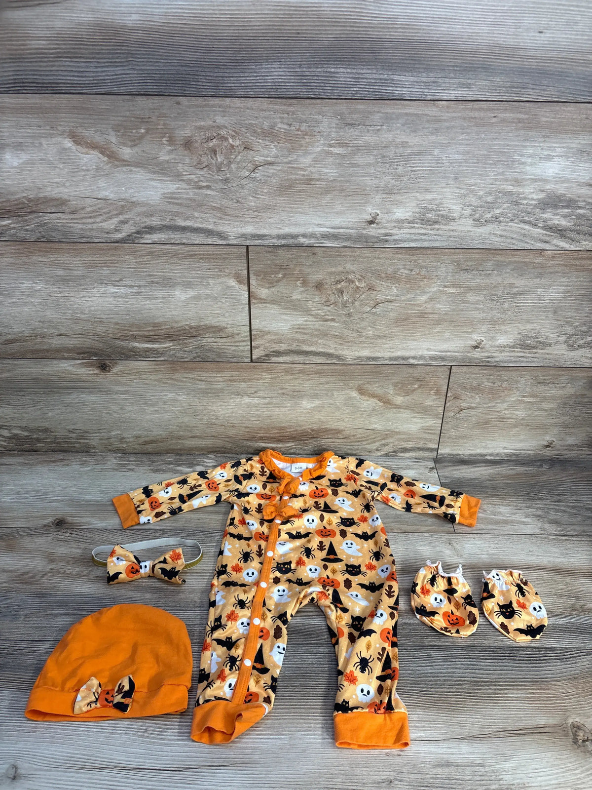 Top-down view of Kucnuzki orange Halloween coverall set with matching mittens, hat, and headband on gray wood flooring