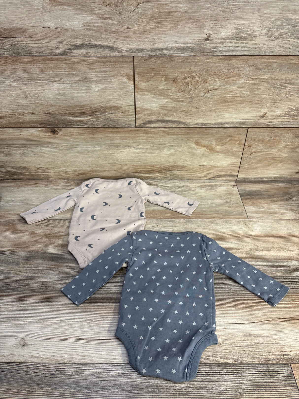Top down of two Cloud Island long sleeve baby bodysuits: grey with stars and light pink with moon print on wood background