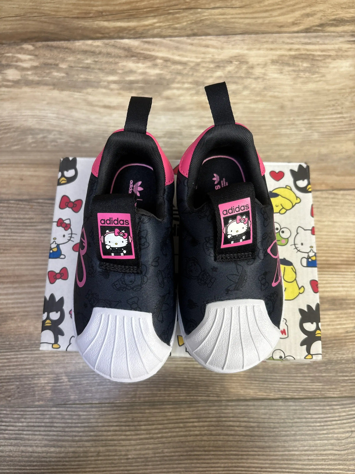 Top view of black Adidas x Hello Kitty Superstar 360 sneakers showing white shell toes and pink Hello Kitty labels on a patterned box