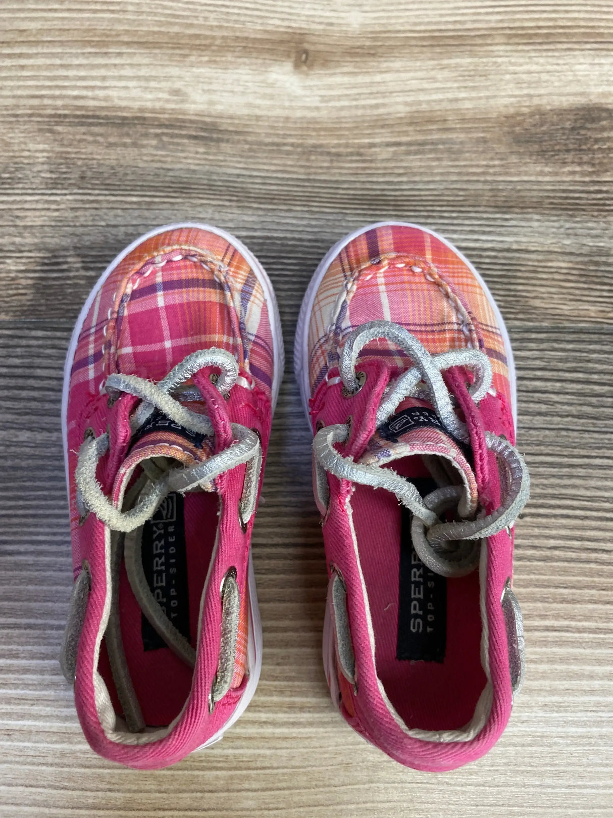 Top-down view of Sperry boat shoes showcasing pink-orange plaid fabric, silver laces, and white soles on a wood surface