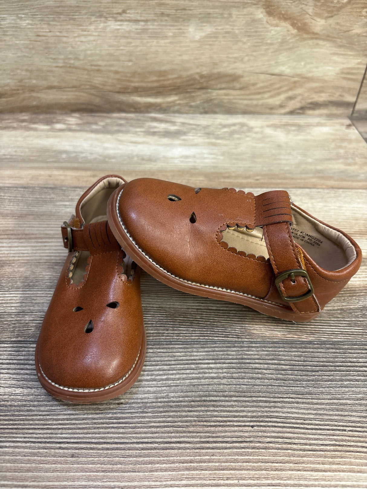 Angled view of brown T-strap Mary Jane shoes (size 9C) showing buckle strap, scalloped cutouts, and stitched sole on wood