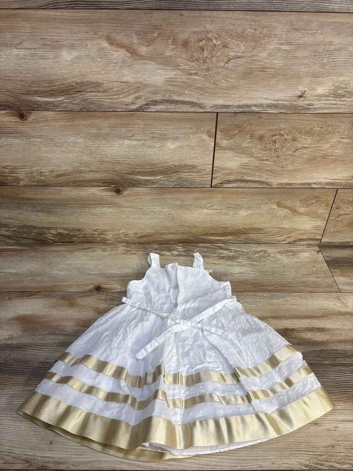 Top-down view of Bonnie Jean white Swiss dot toddler dress with gold ribbon hem and tied waist sash on wood background
