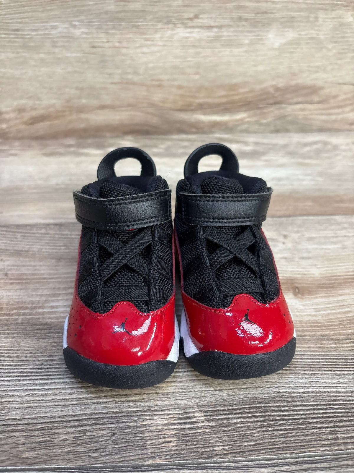 Front view of toddler Jordan 6 Rings 'Fitness Red' sneakers showing glossy red patent toe, black mesh upper, velcro strap on woodgrain background
