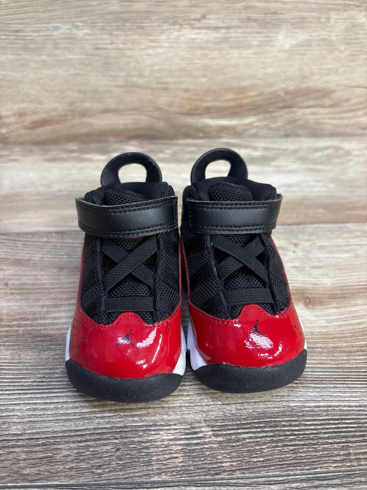 Front view of toddler Jordan 6 Rings 'Fitness Red' sneakers showing glossy red patent toe, black mesh upper, velcro strap on woodgrain background