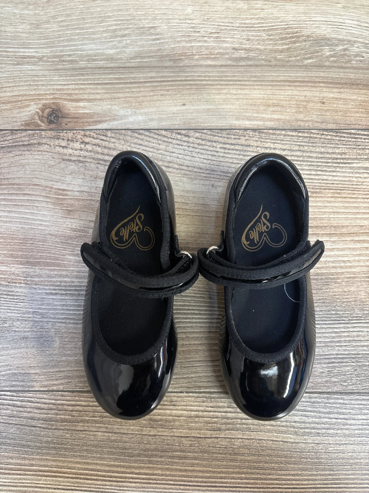 Top-down view of Stelle girls tap dance shoes, size 7C, showing black patent finish and Velcro tap straps on wood floor