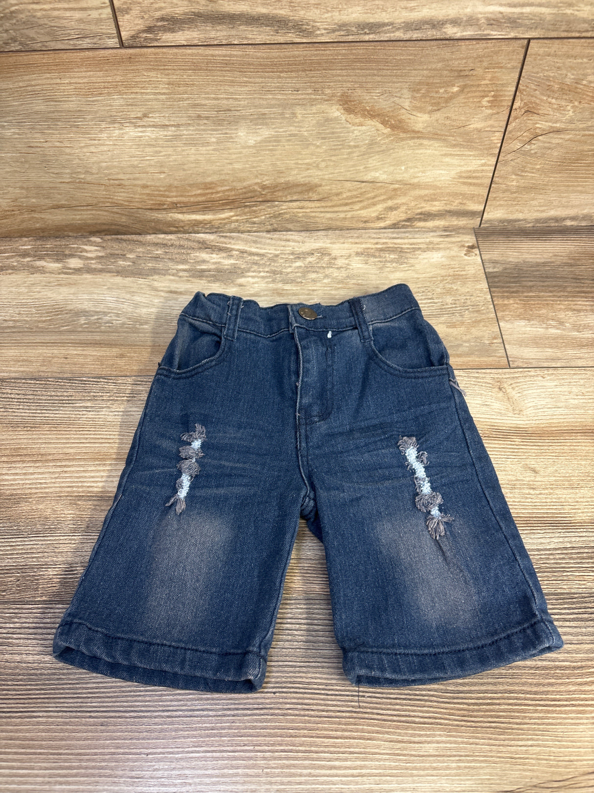 Front view of size 2T blue distressed denim toddler shorts showing ripped knee details and elastic waistband on wood floor background