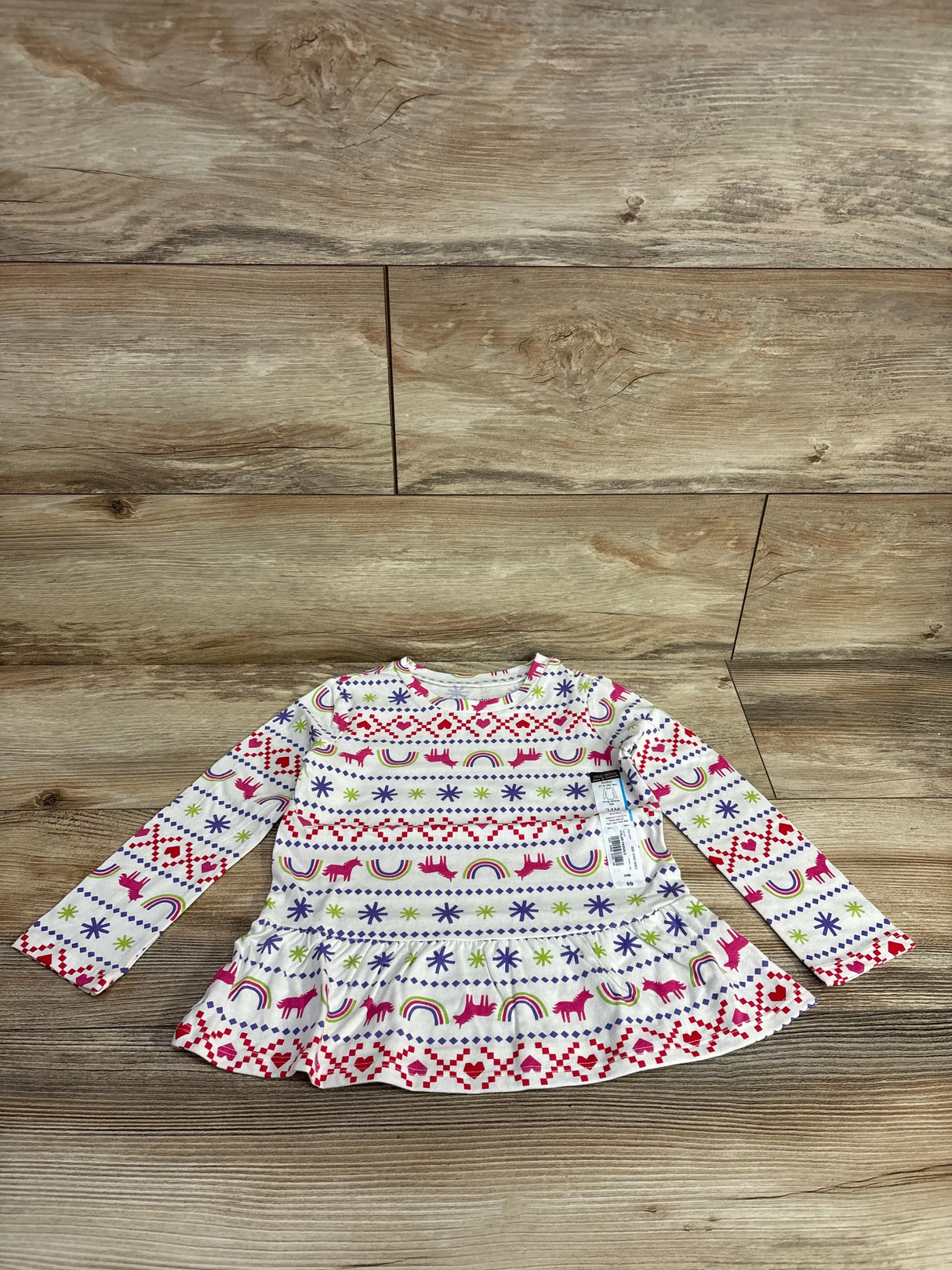 Front view of white Okie Dokie peplum toddler shirt showcasing unicorn, rainbow, heart and star Fair Isle print on wood floor