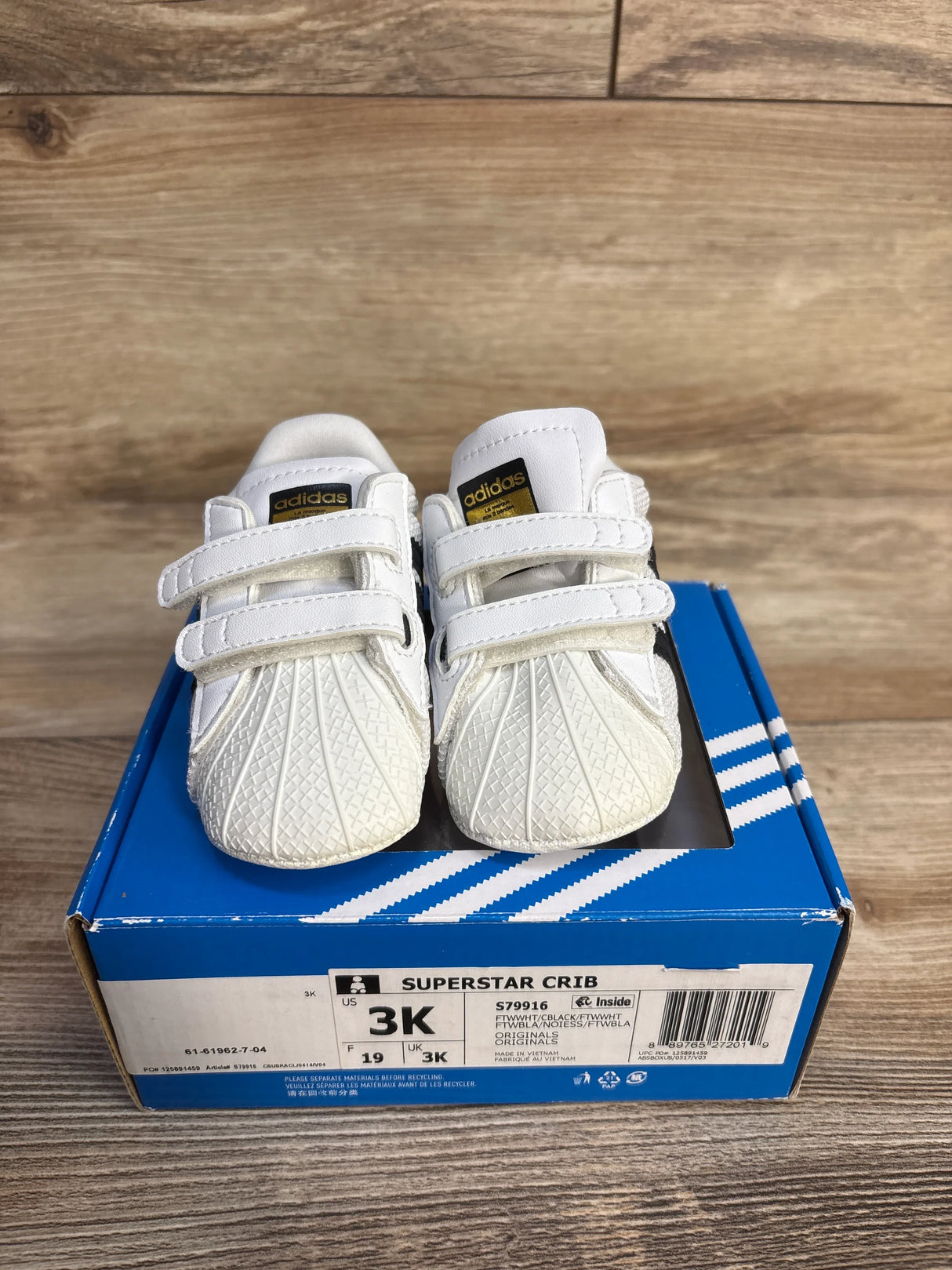 Adidas Superstar Crib infant shoes white with Velcro straps on blue box, wood background