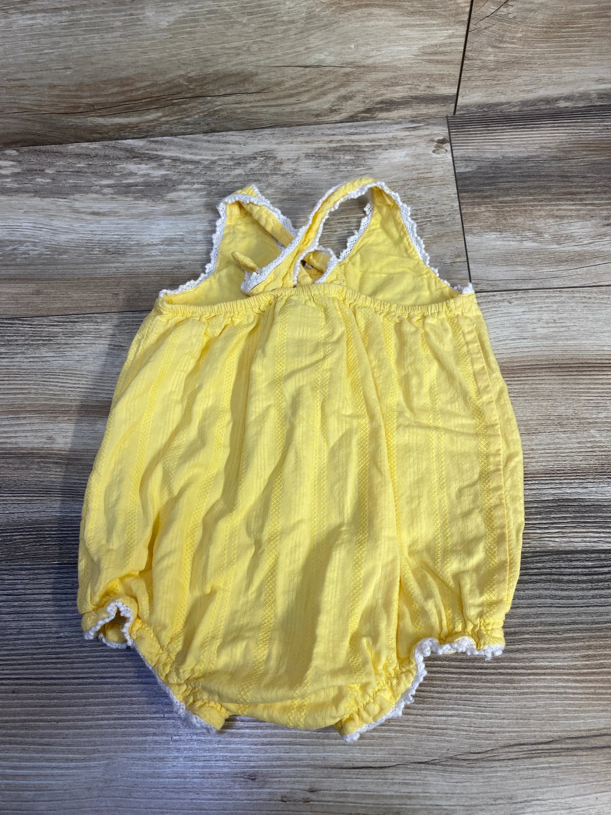 Flat lay of yellow baby romper with crisscross back straps, white lace trim, and elastic leg openings on wood background