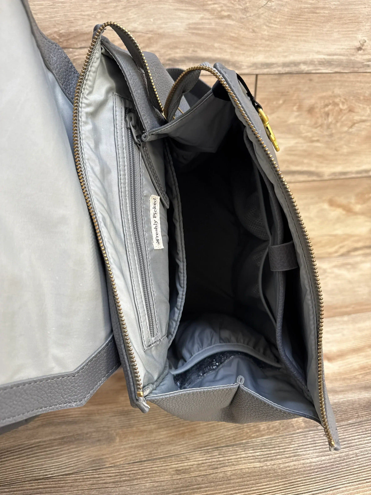 Top-down view of Freshly Picked Stone diaper bag II showing open interior with zip and mesh pockets on wood floor