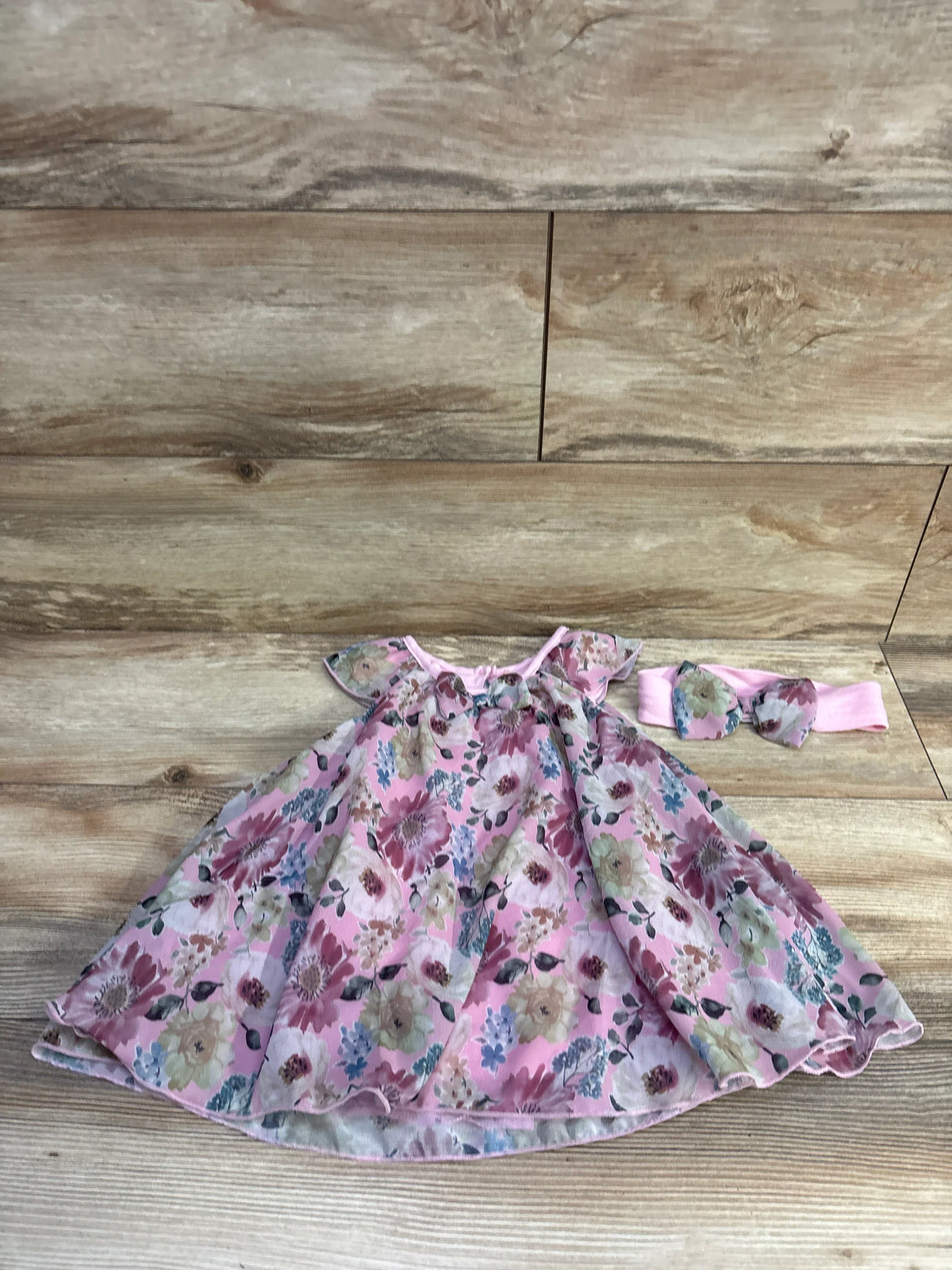 Flat lay of pink floral baby bodysuit dress and matching bow headband showing flutter sleeves and gathered skirt on wood background