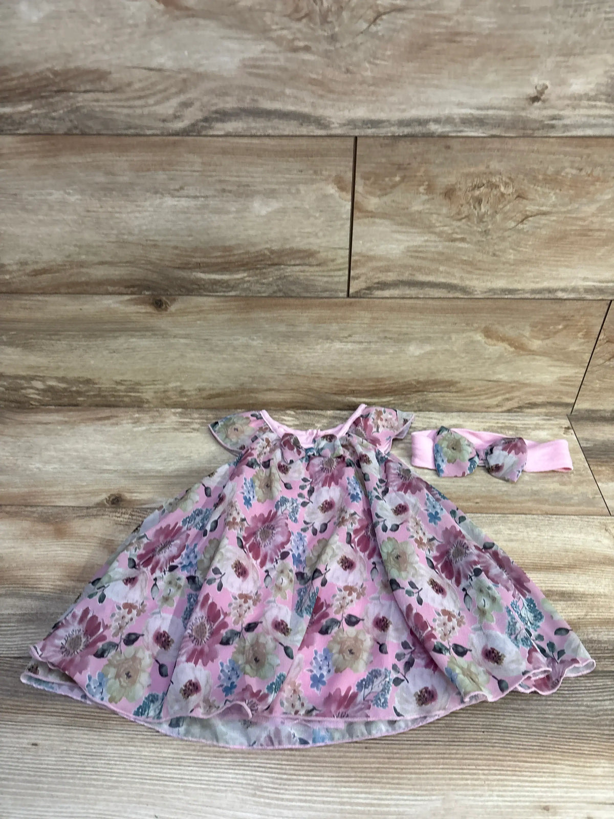 Flat lay of pink floral baby bodysuit dress and matching bow headband showing flutter sleeves and gathered skirt on wood background