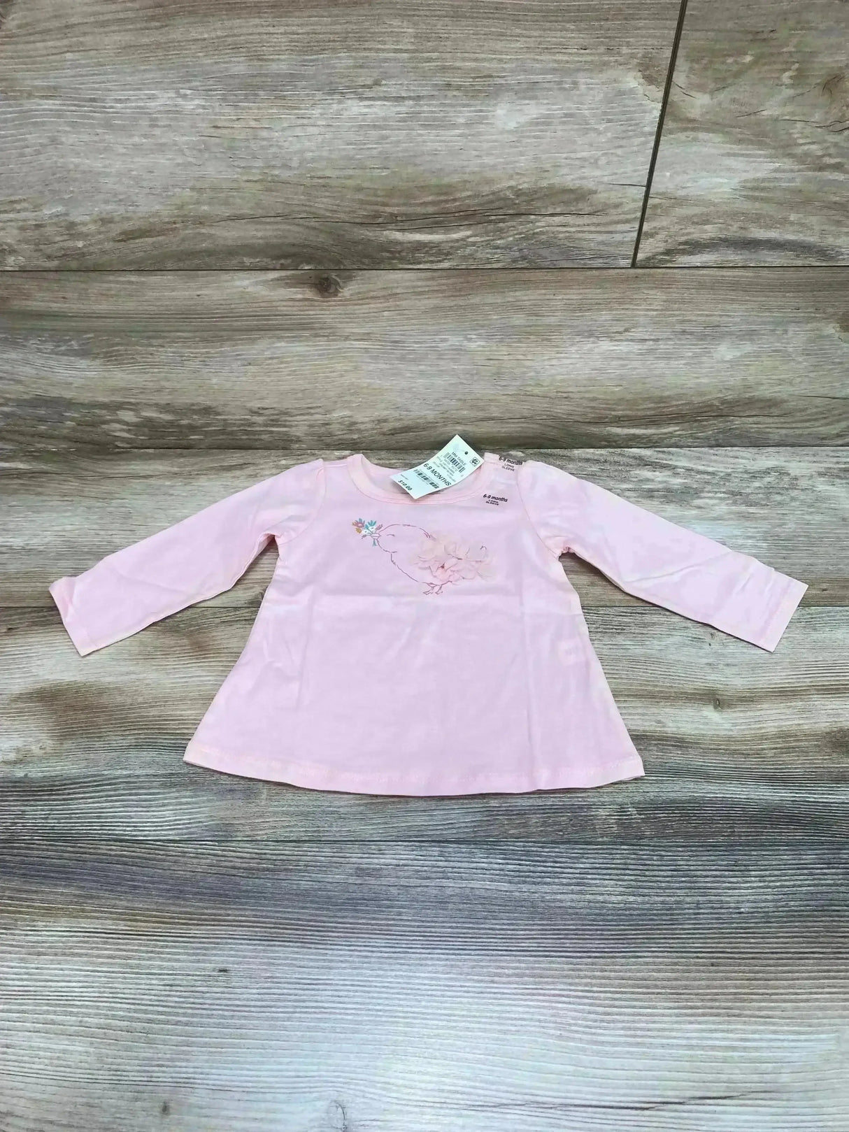 Front flat lay of pink long-sleeve baby tunic showing embroidered bird with floral accents and attached size tag on wood background
