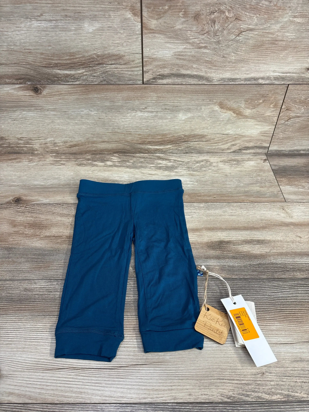 Top-down view of Kickee Kids peacock casual baby pants showing folded cuffs and attached brand tags on wood floor