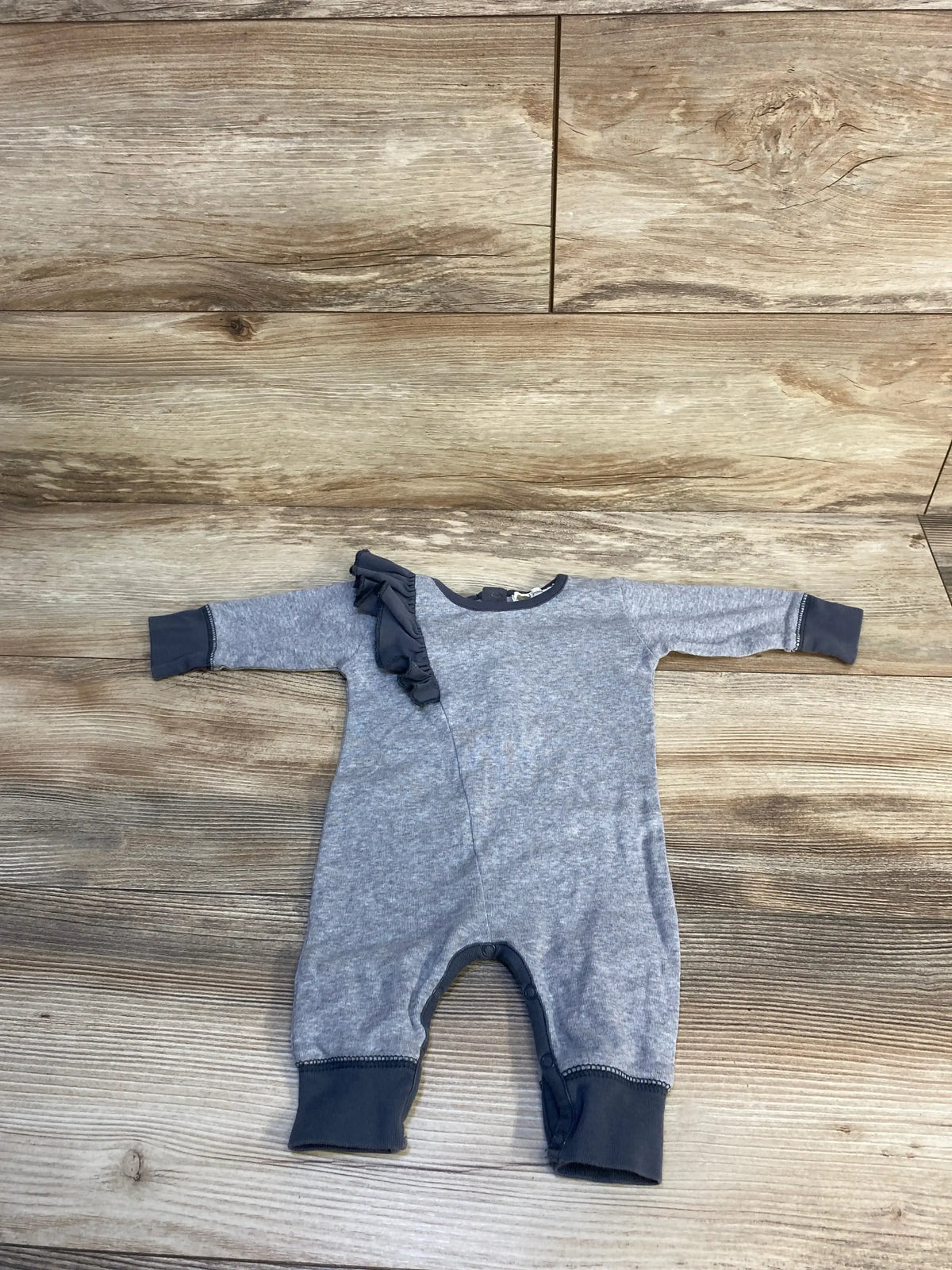 Flat lay of Monica + Andy grey newborn coverall showing left shoulder ruffle and snap leg closures on wooden floor