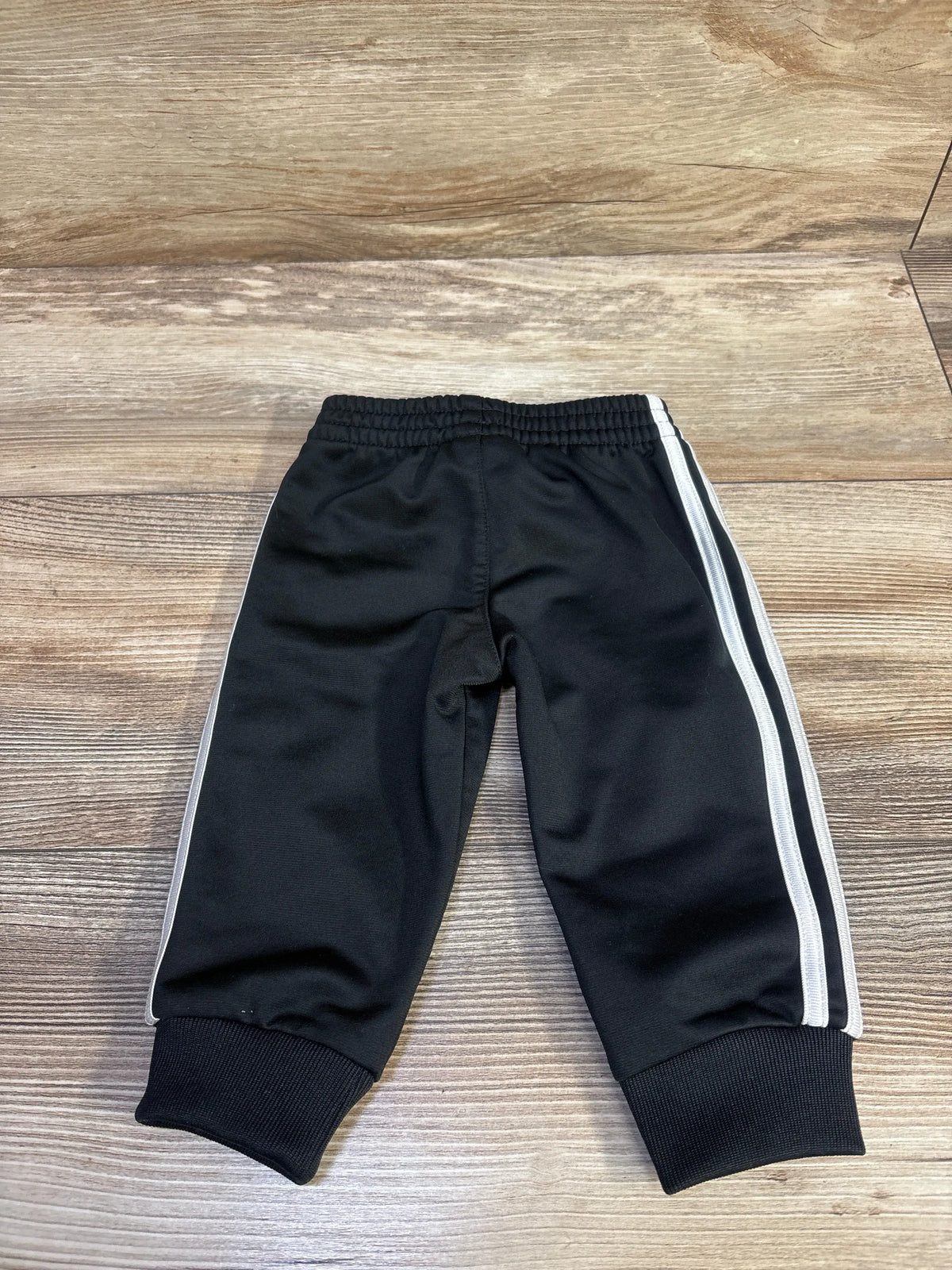 Front view of Adidas black joggers in size 6 months showcasing white side stripes and ribbed cuffs on a wood-look floor