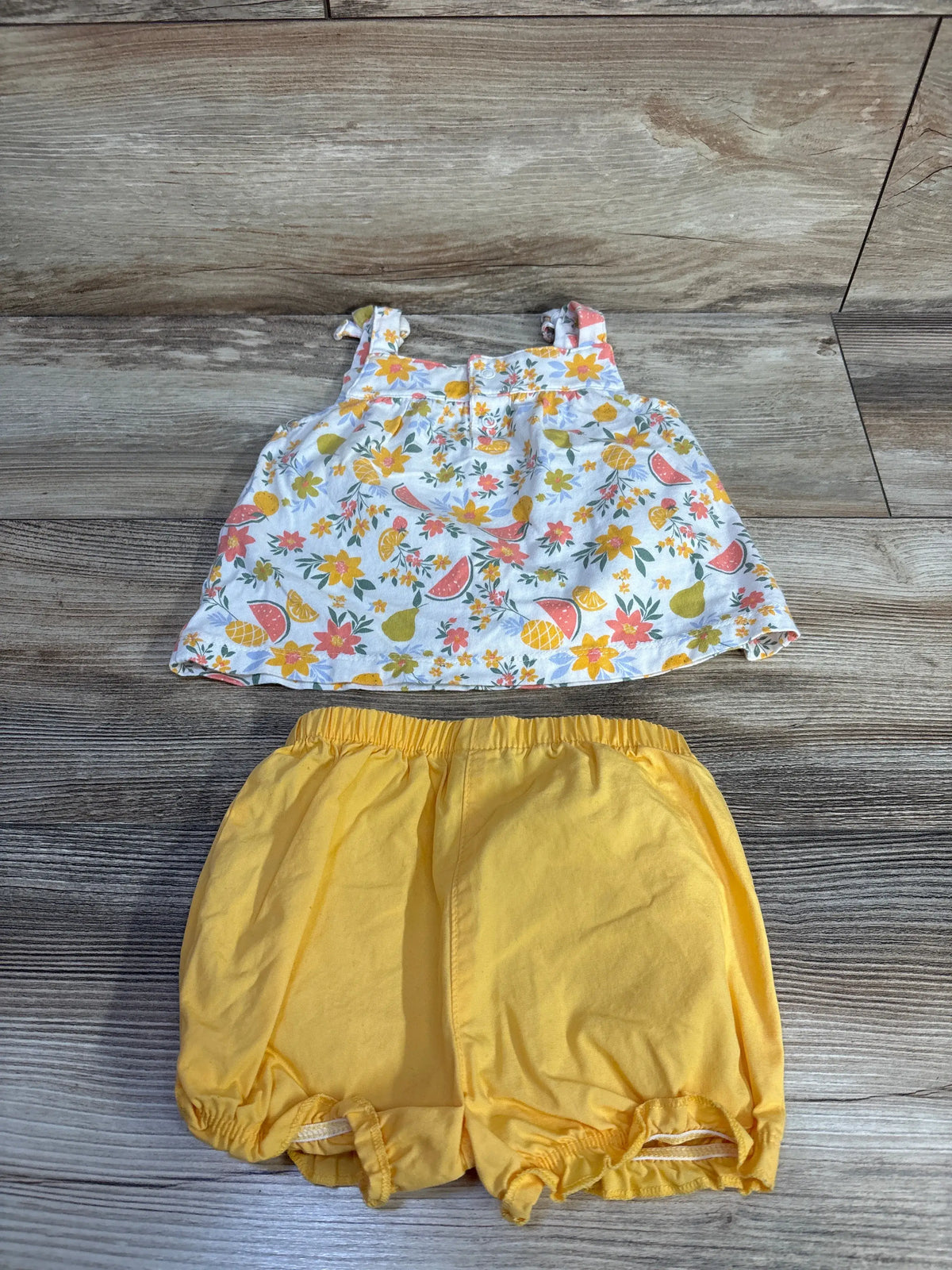 Flat lay of Just One You 2-piece tank top and shorts set showcasing a floral tank and yellow shorts on wood flooring
