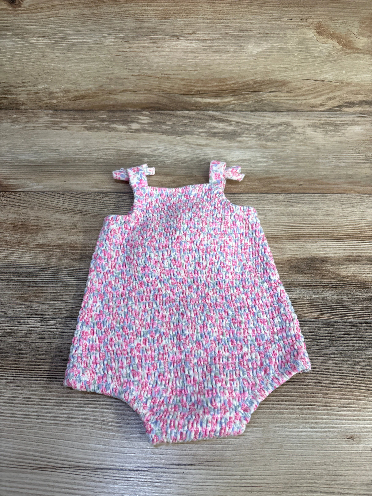 Front view of pink smocked bubble romper (3-6 months) showing smocked texture, tied shoulder straps, and elastic leg openings on a wood surface