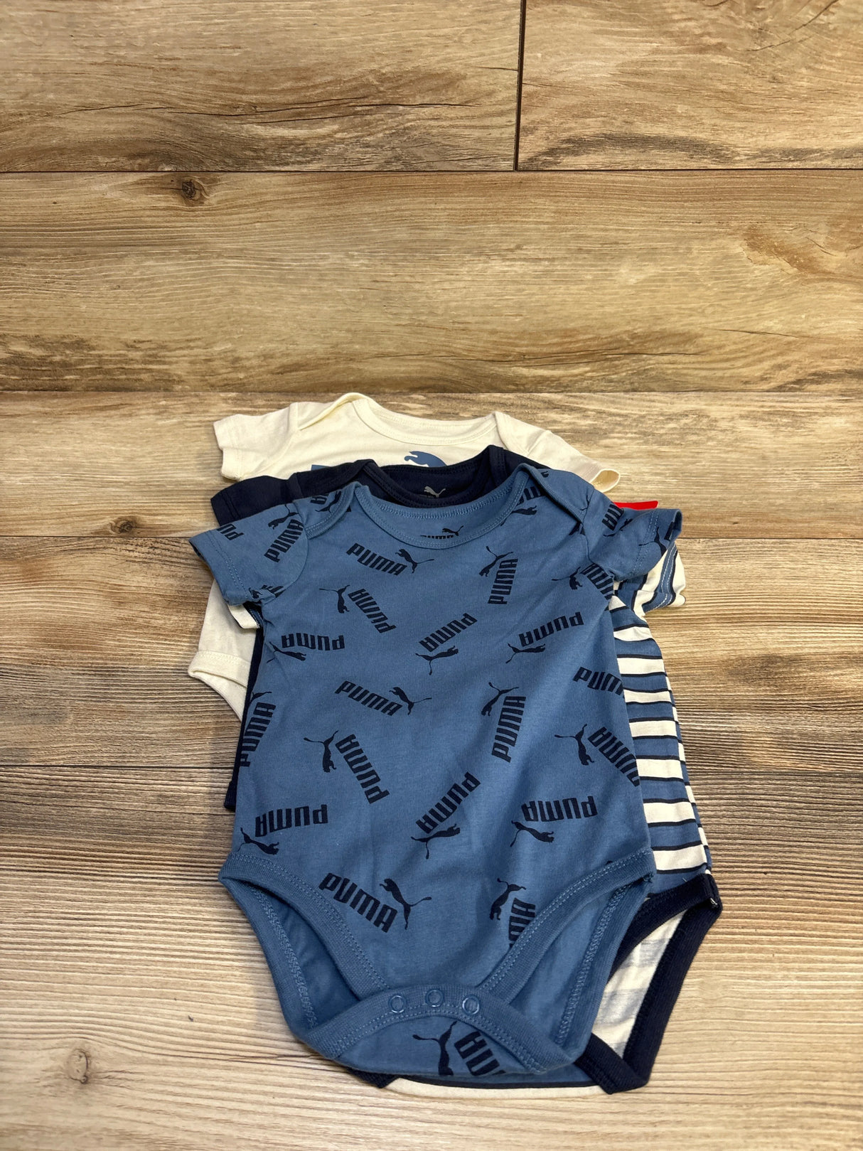 Top view of a 4-pack of Puma baby bodysuits showing a blue bodysuit with repeated Puma logos and navy, cream, and striped suits on a wood background