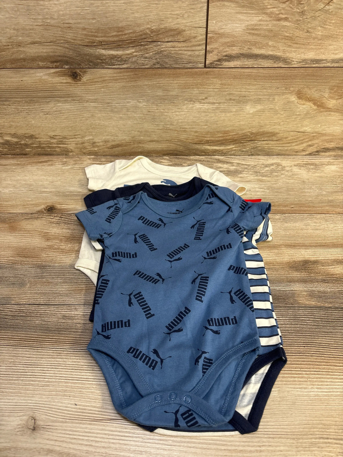 Top view of a 4-pack of Puma baby bodysuits showing a blue bodysuit with repeated Puma logos and navy, cream, and striped suits on a wood background