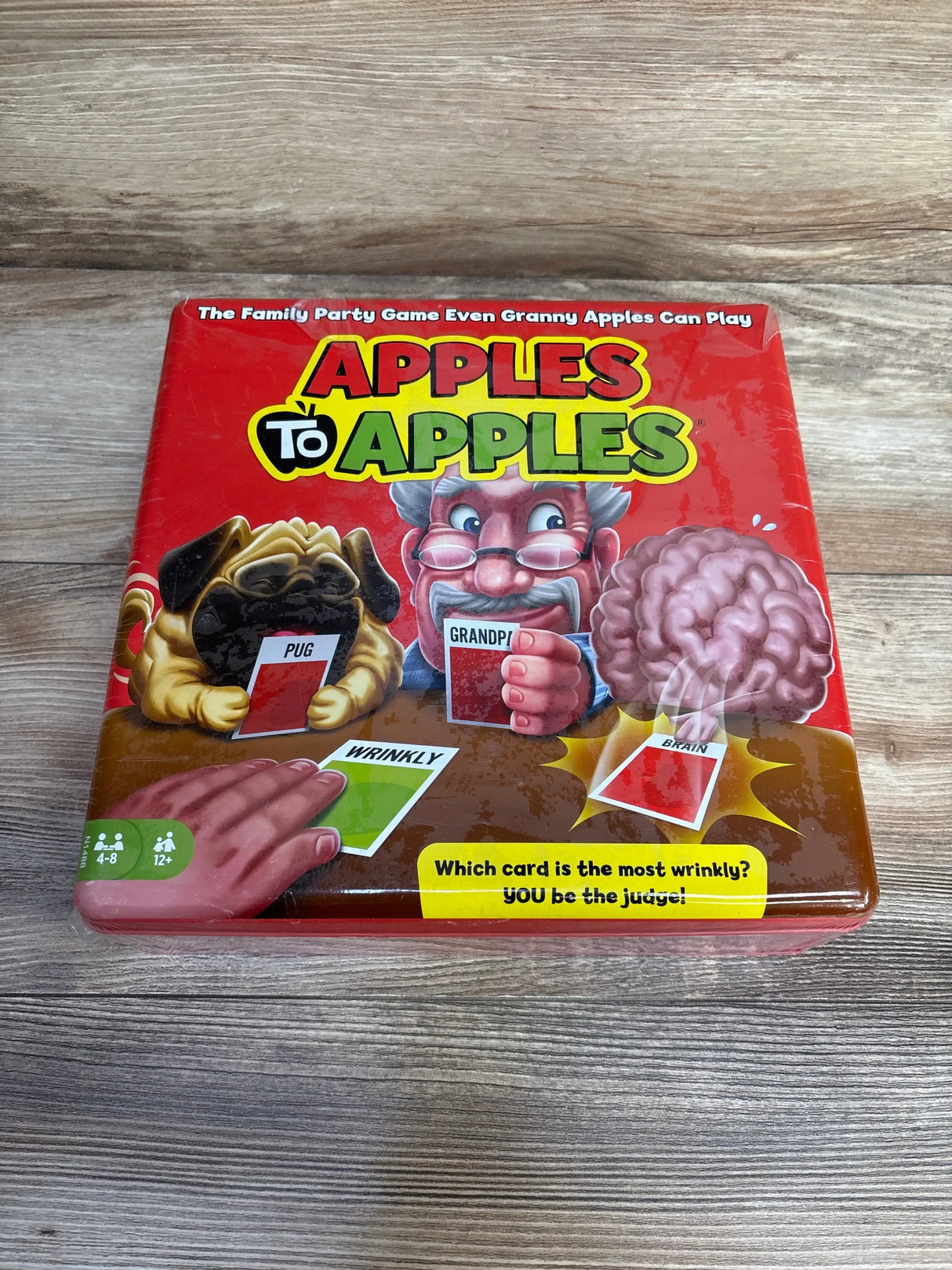 Front of Apples to Apples game tin showing title Apples to Apples, characters and cards on wooden tabletop