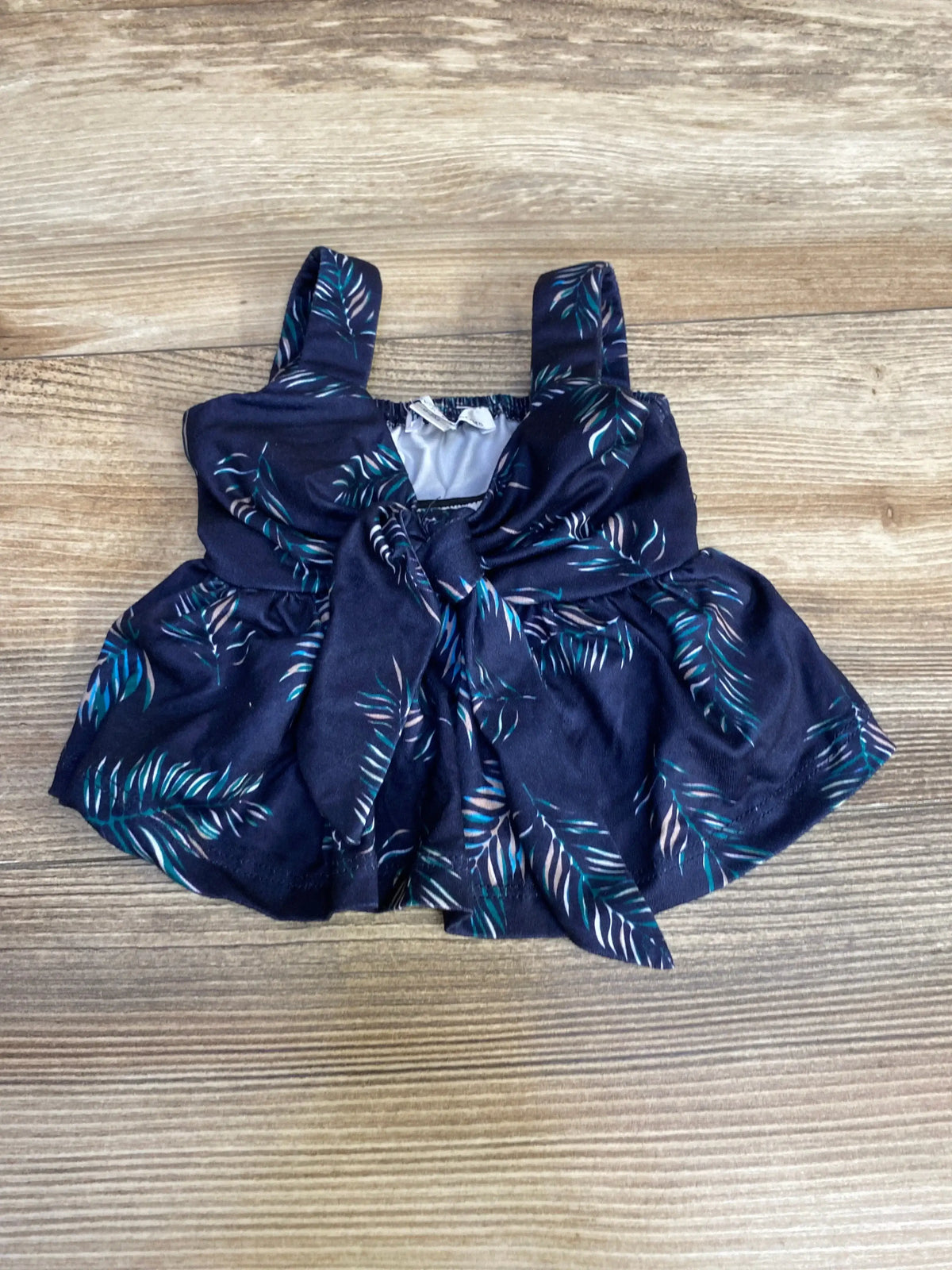 Top-down of Bailey's Blossoms navy sleeveless baby top with front tie bow, teal leaf print, gathered waist and straps on wood background