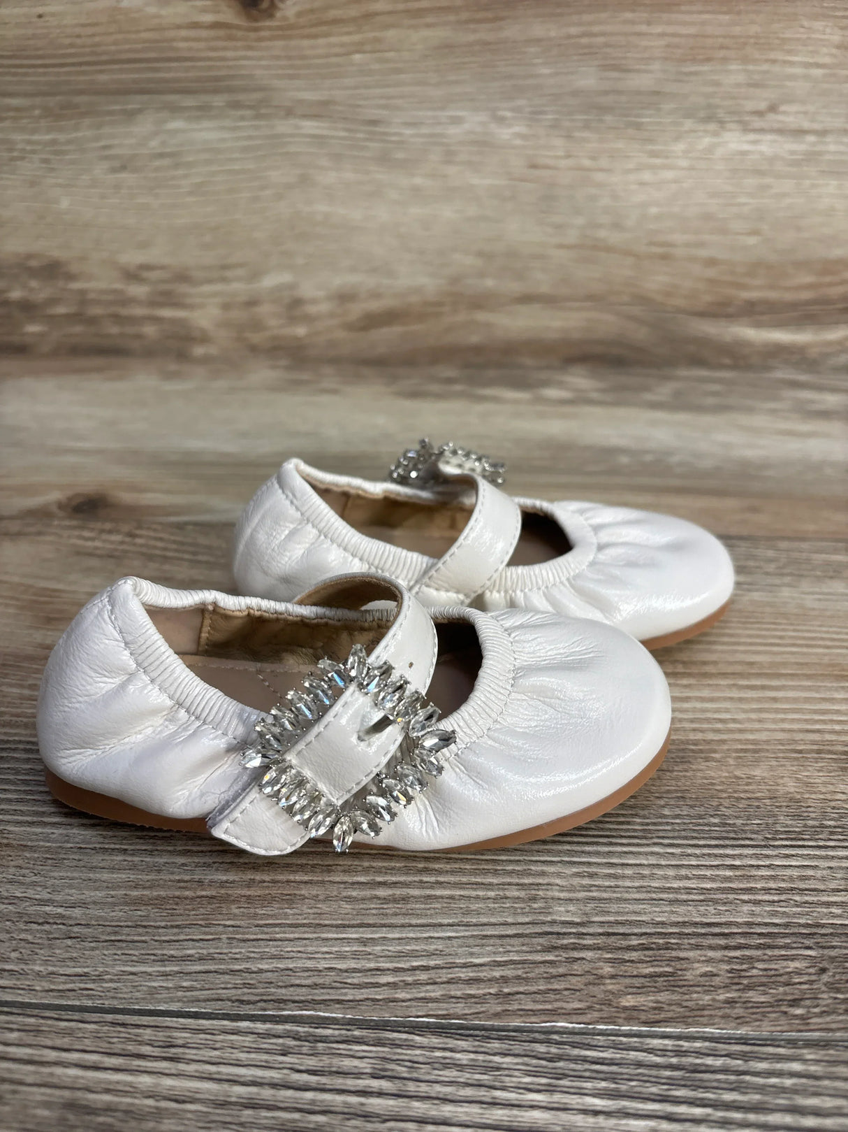 White leather baby shoes with crystal buckle on wooden floor, soft sole and elastic trim