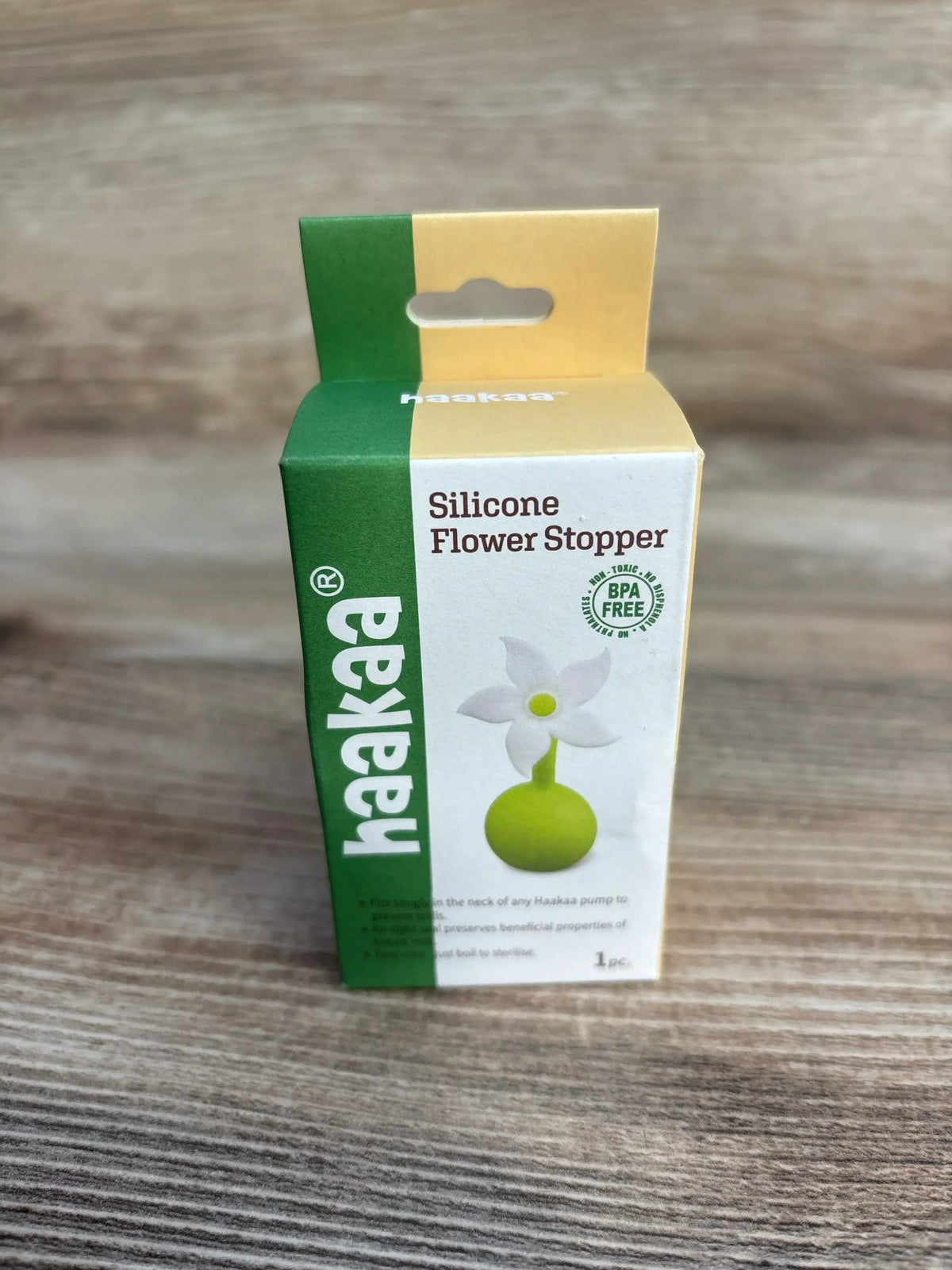 Front view of NEW haakaa Silicone Flower Stopper in green and yellow BPA free box on wood plank surface