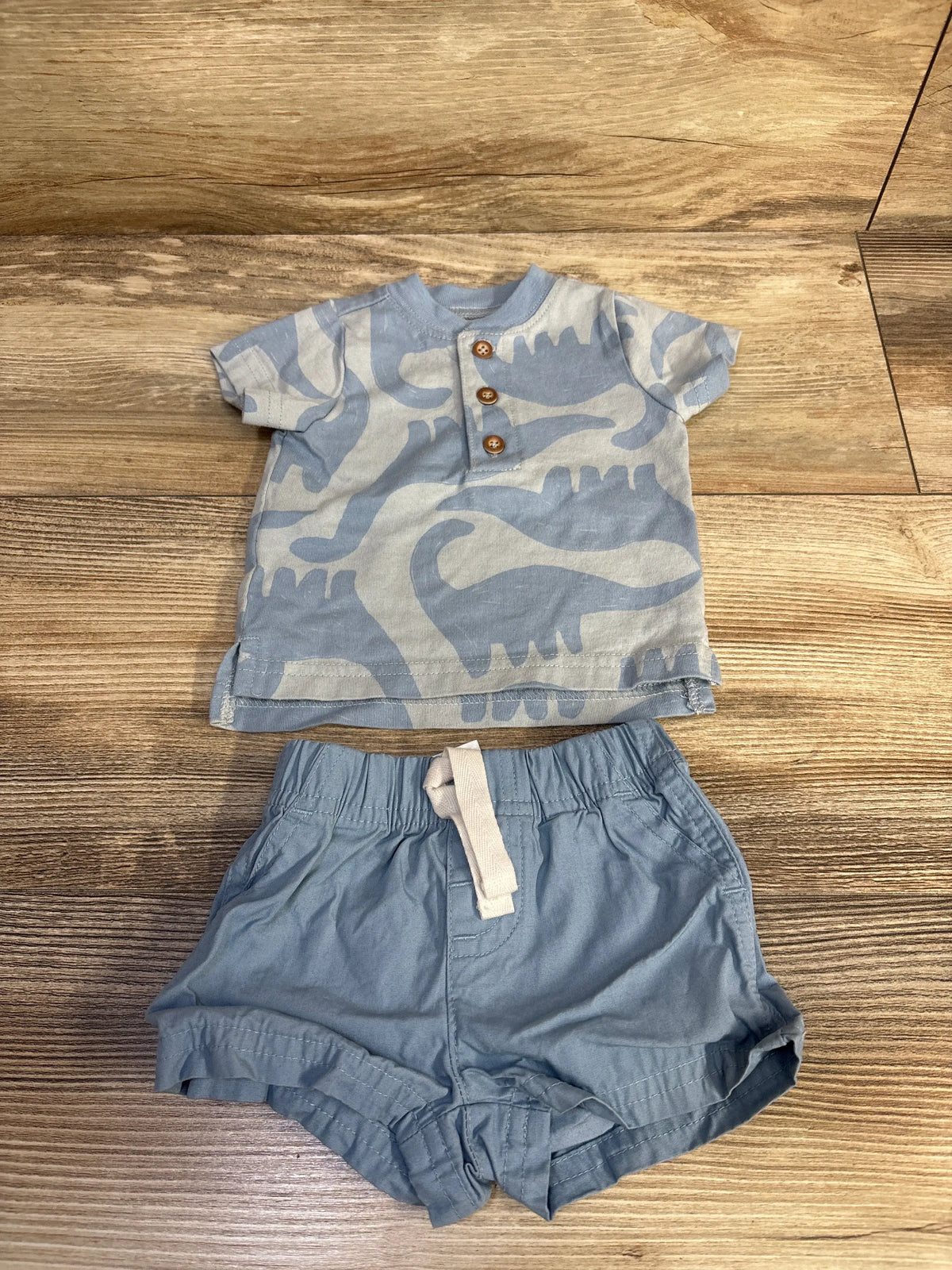 Front view of Just One You henley shirt and shorts set showcasing a blue dinosaur-patterned shirt on a wood surface