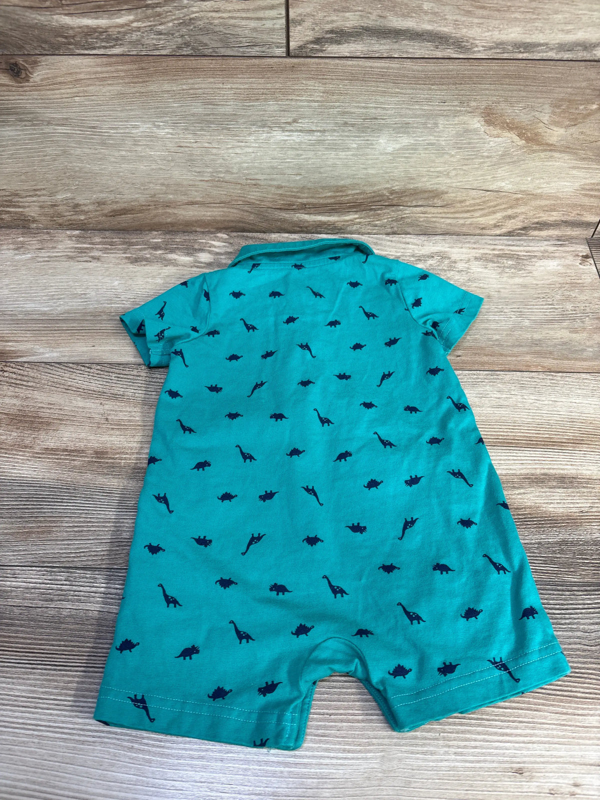 Front view of green dinosaur-print polo shortie romper with collared short sleeves on a wood-grain background