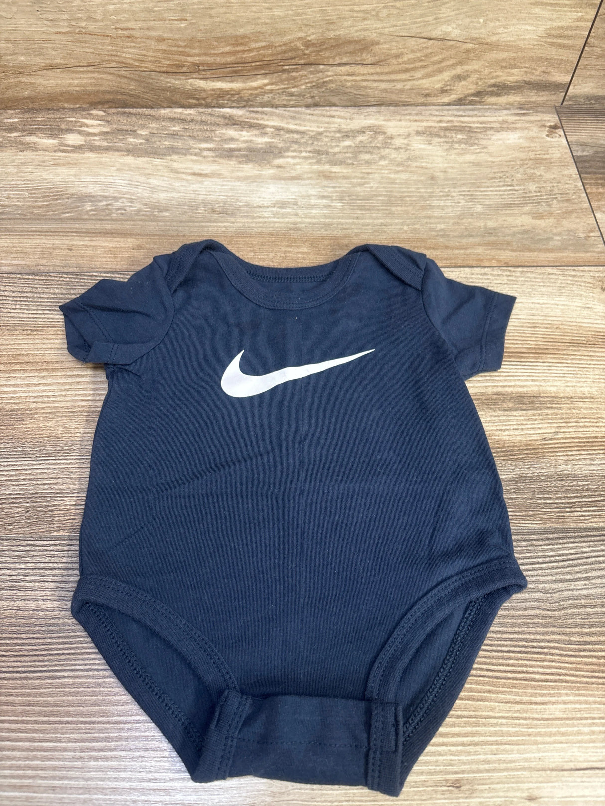 Front view of navy Nike baby bodysuit showcasing white swoosh logo, size 3M, on wood floor background