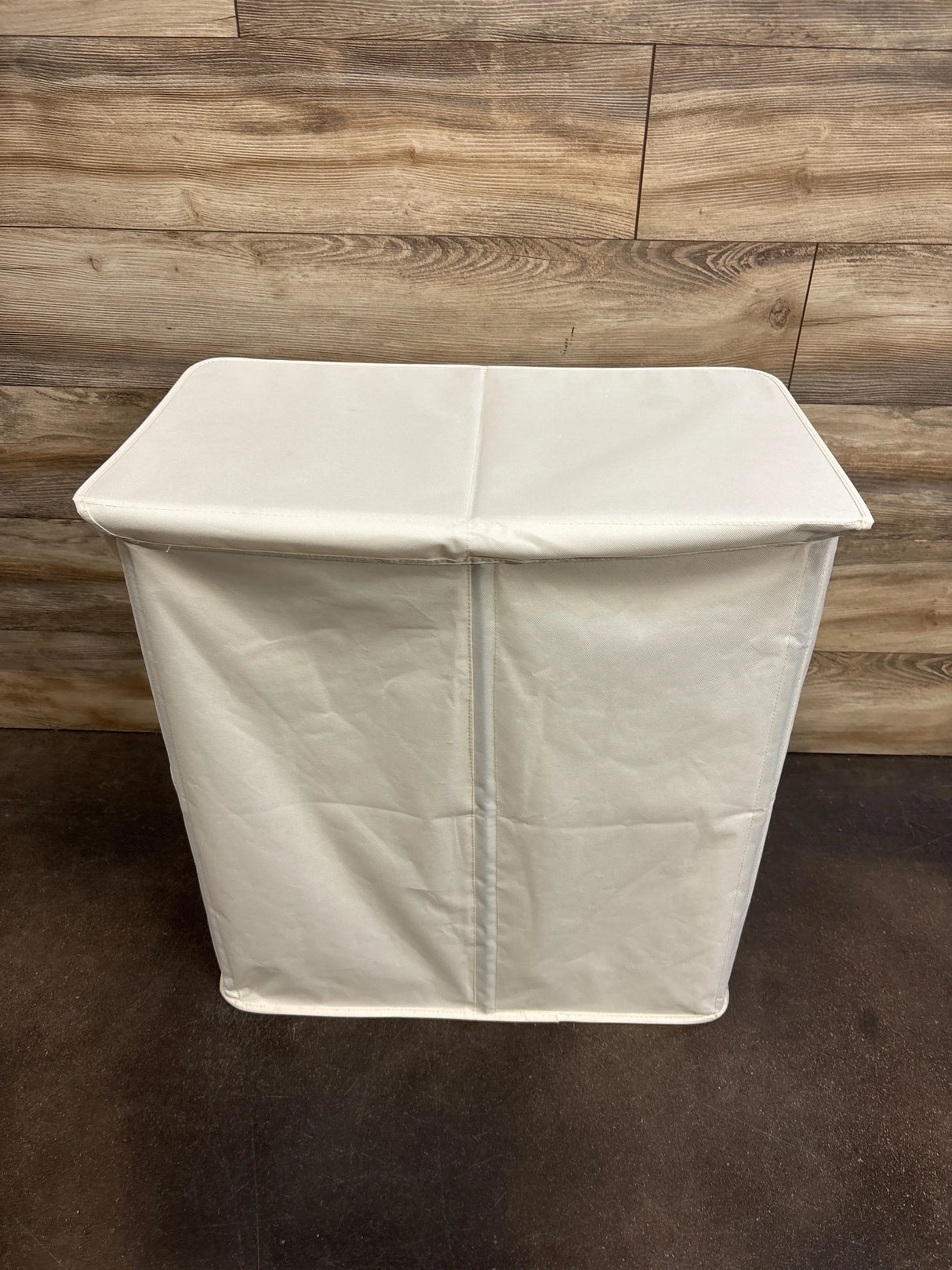 Angled front view of white double laundry hamper with closed lid showing center divider and fabric against wood wall
