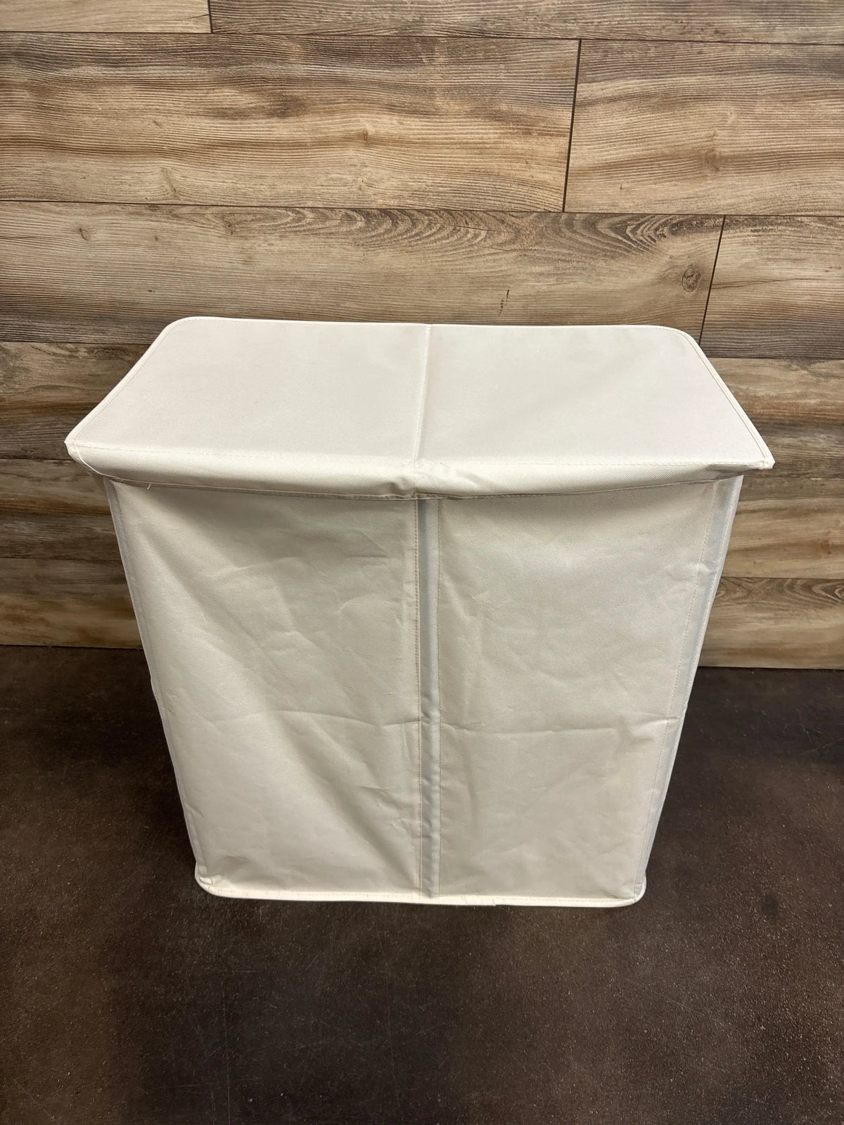Angled front view of white double laundry hamper with closed lid showing center divider and fabric against wood wall