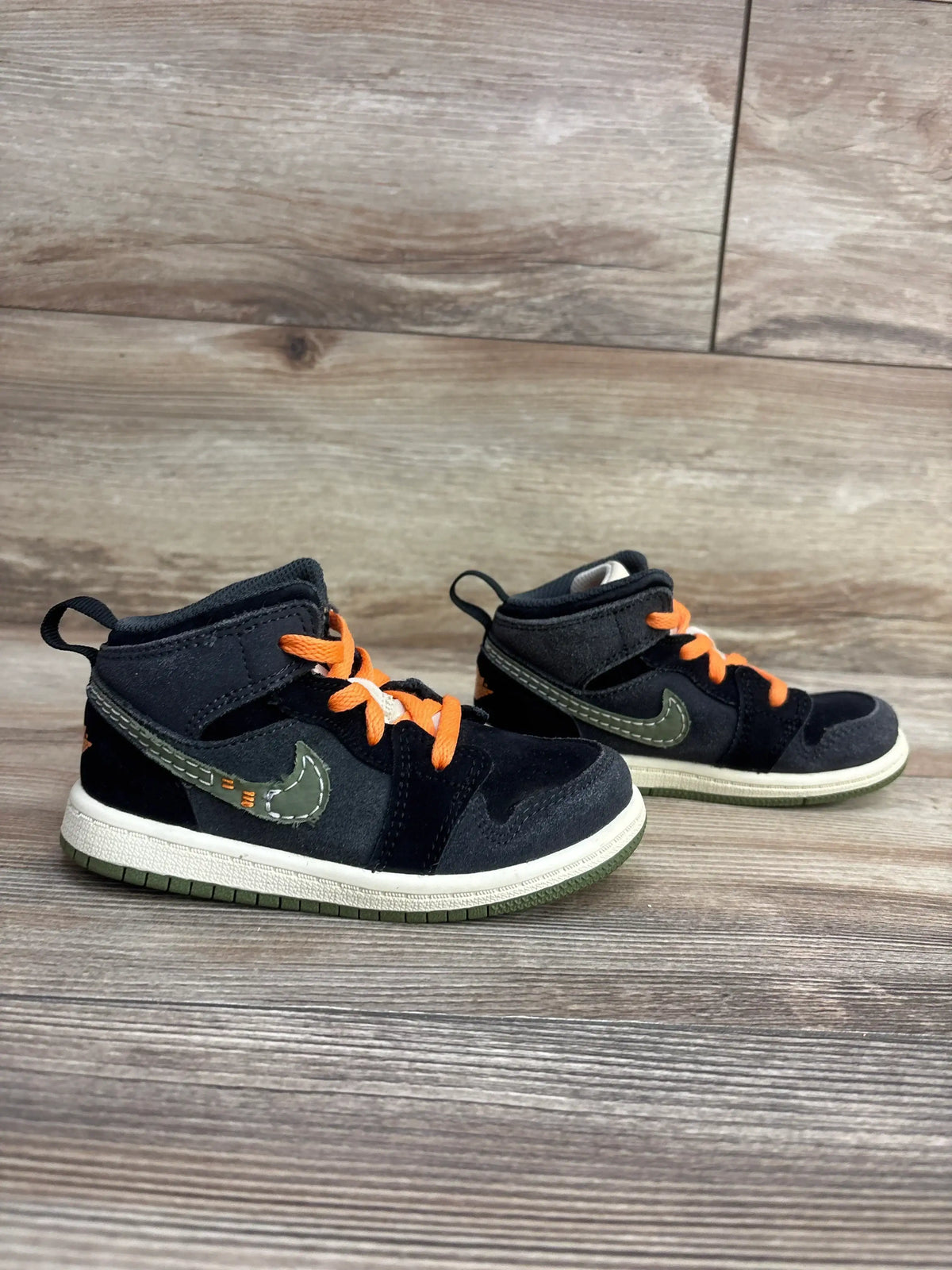 Side view of Air Jordan 1 Mid toddler sneakers in anthracite and light olive showcasing orange laces and olive swoosh on wood background